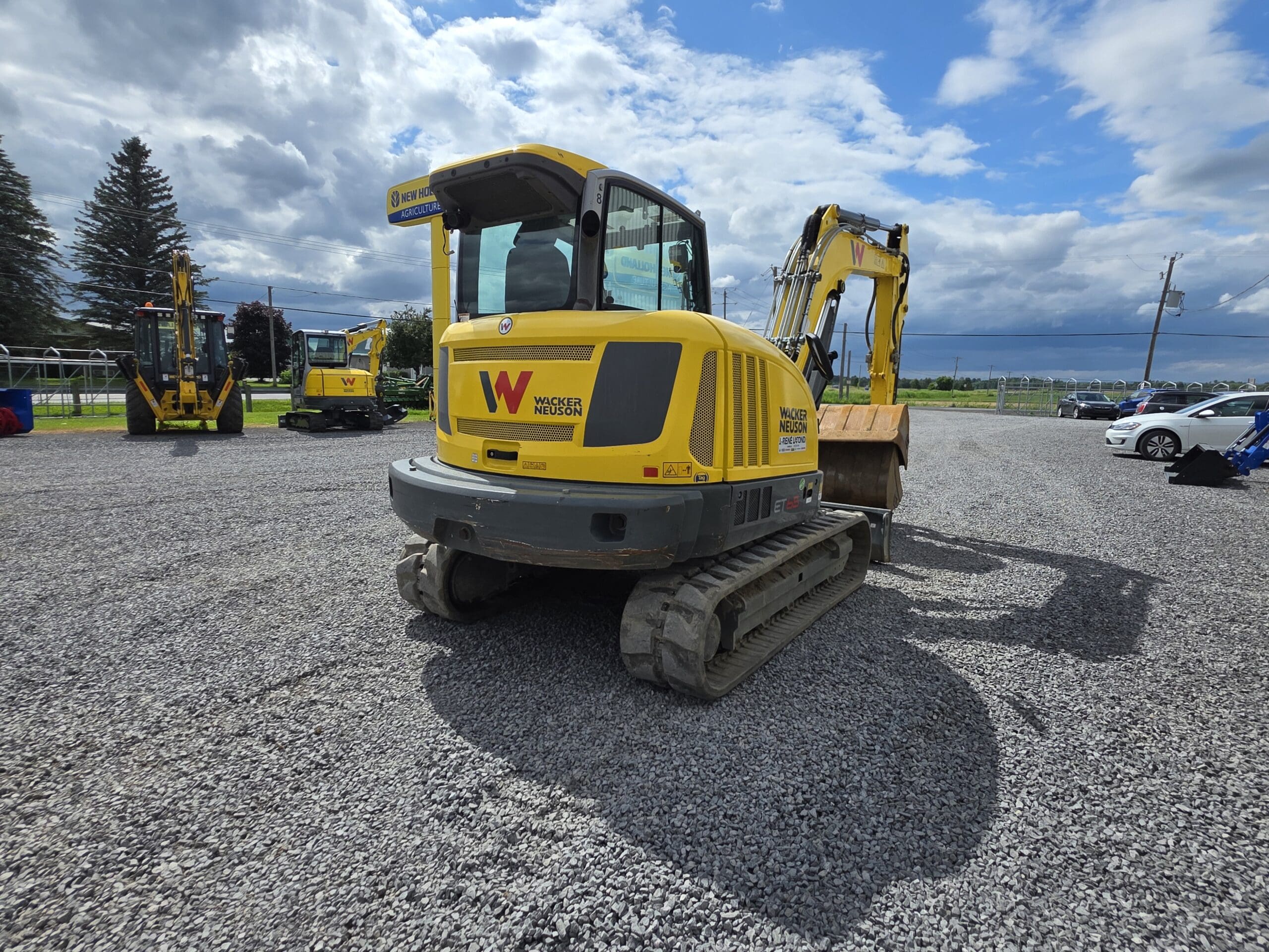 Une pelleteuse Wacker Neuson jaune est posée sur un terrain en gravier sous un ciel partiellement nuageux, avec une autre pelleteuse, des voitures et des arbres visibles à l'arrière-plan.