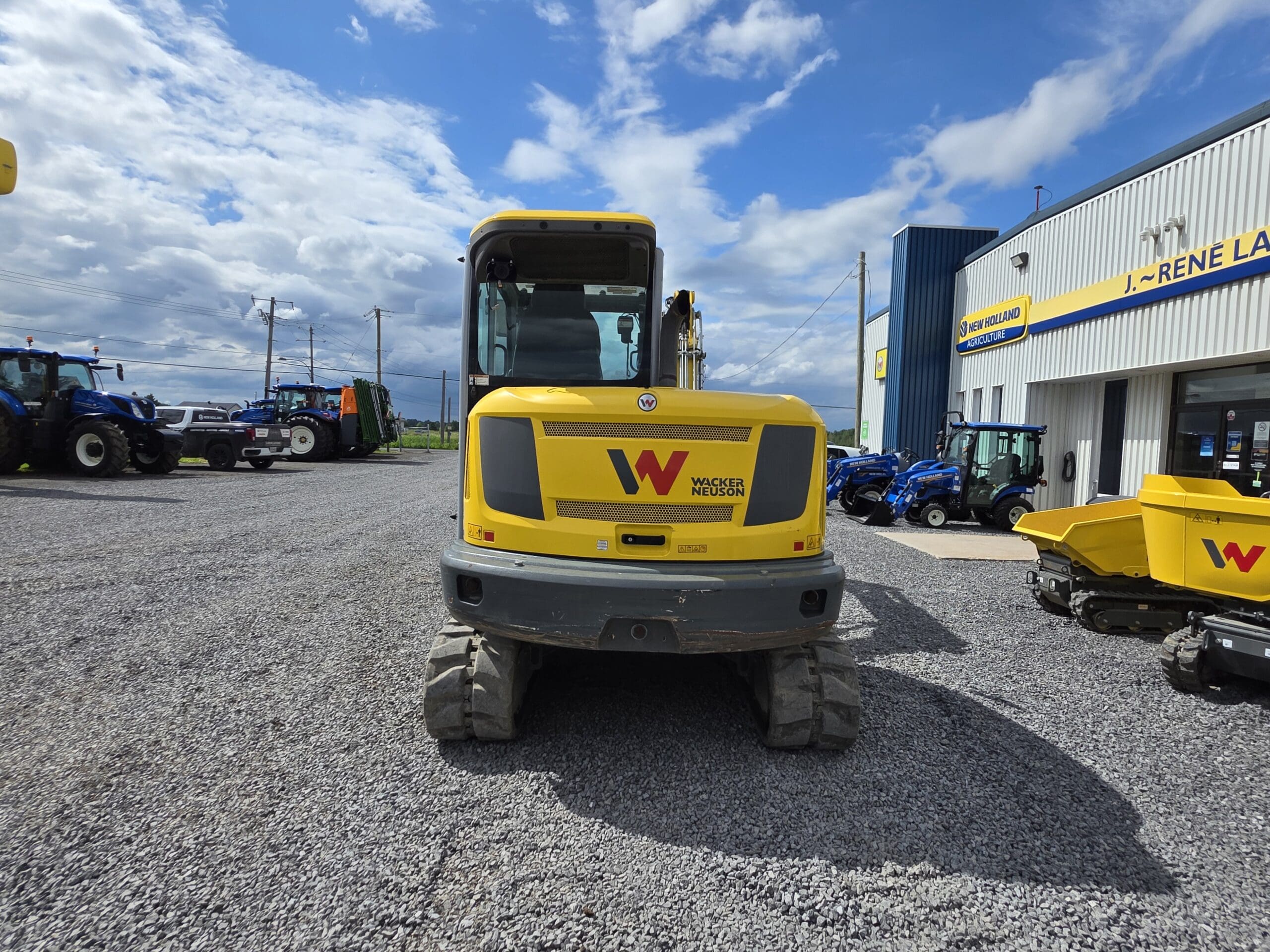 Un véhicule de construction Wacker Neuson jaune est garé sur un terrain en gravier à l'extérieur d'une concession. D'autres machines et équipements sont visibles à l'arrière-plan sous un ciel partiellement nuageux.