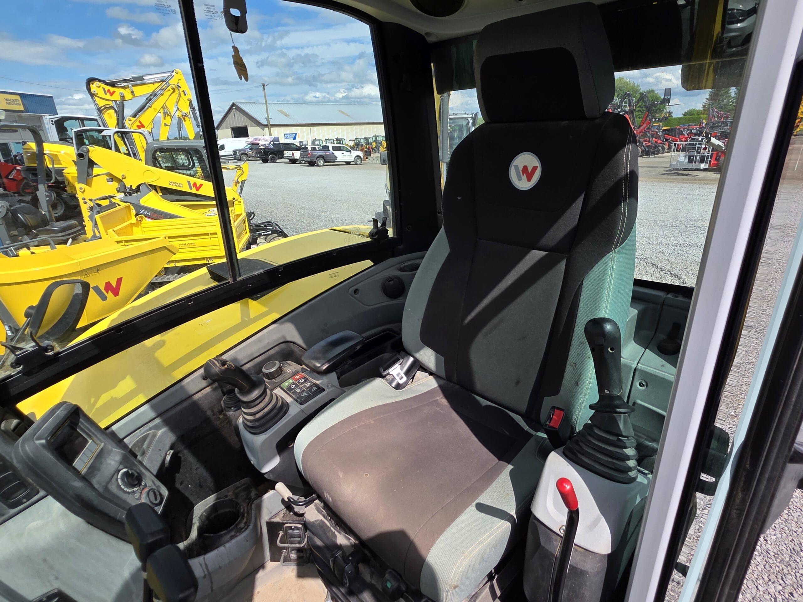 Vue du siège du conducteur et des commandes à l'intérieur de la cabine d'un véhicule de construction jaune, avec des manettes, des boutons et des fenêtres montrant d'autres équipements et machines à l'extérieur par une journée ensoleillée.