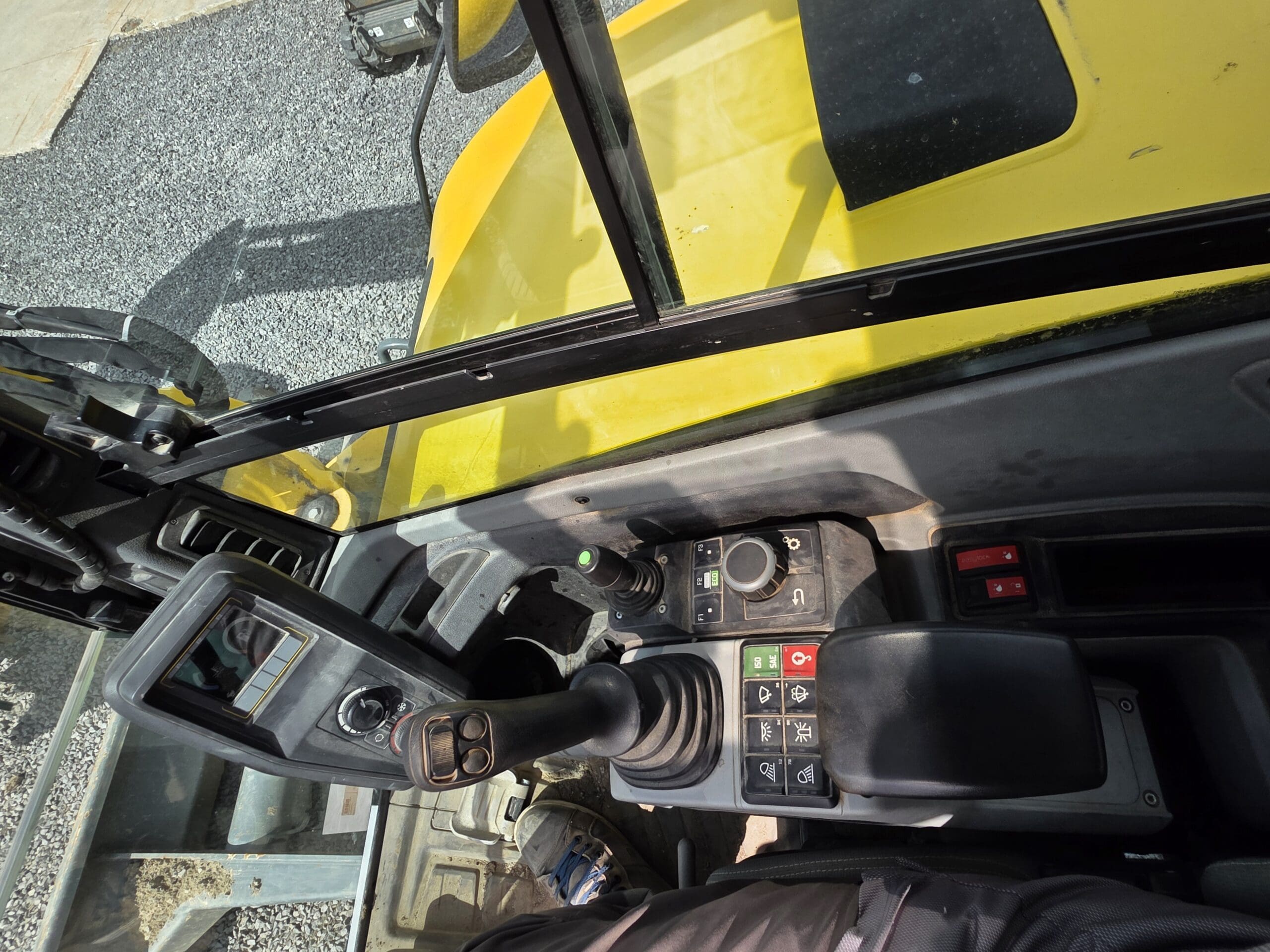 Vue de l'intérieur de la cabine d'un véhicule de chantier, montrant les panneaux de commande, le joystick, les boutons et une partie de l'extérieur jaune. Le sol à l'extérieur est recouvert de gravier.