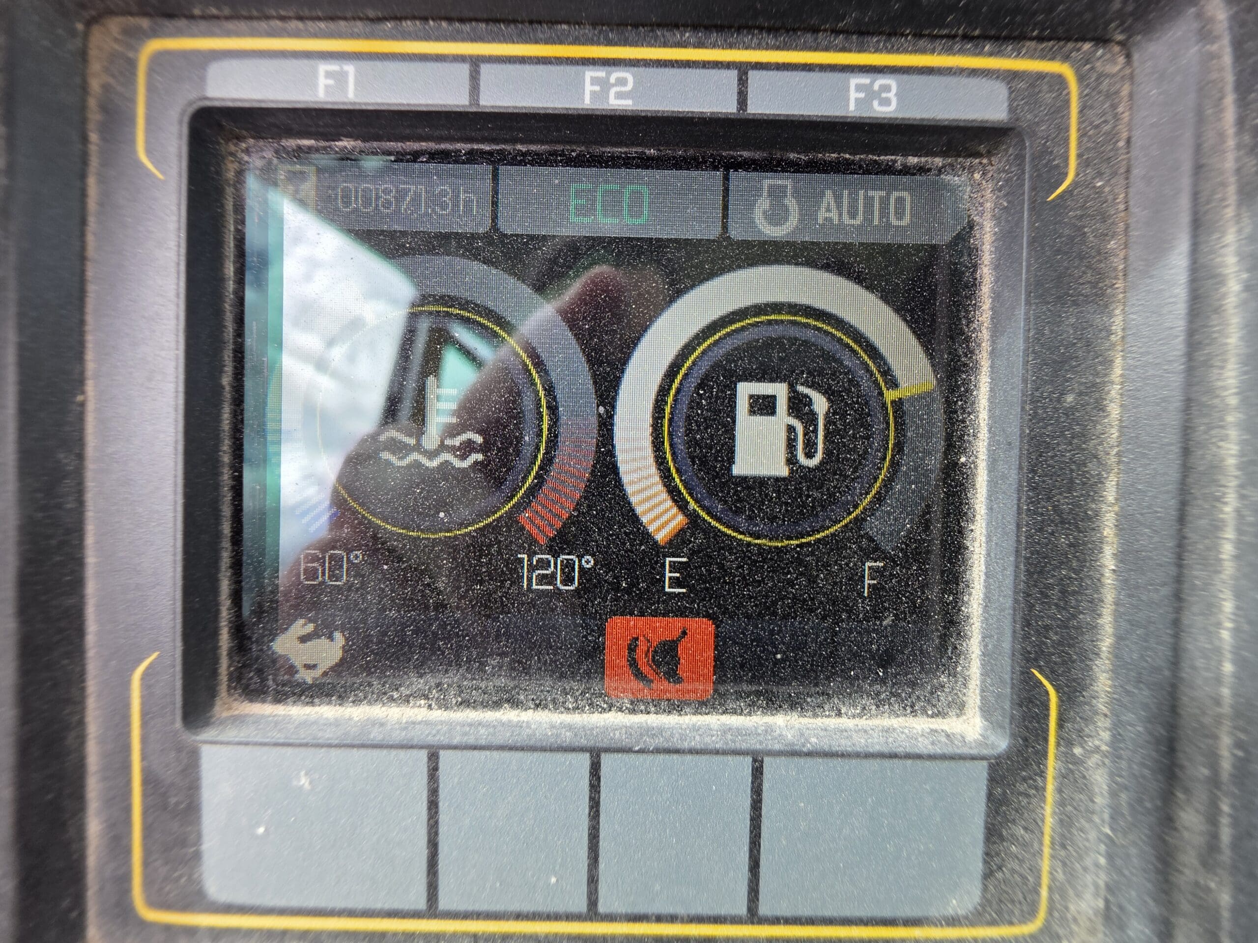 Gros plan sur l'écran du tableau de bord d'un véhicule poussiéreux indiquant la température du moteur, la jauge de carburant presque vide, le mode ECO et les réglages automatiques. L'écran affiche également le compteur horaire à 00871.3h.