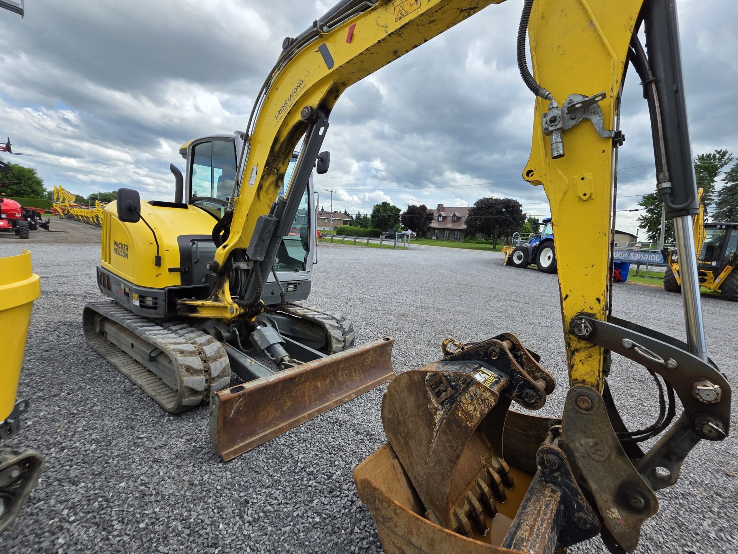 Une pelleteuse compacte jaune munie d'un godet est garée sur un terrain en gravier sous un ciel nuageux, d'autres véhicules de construction étant visibles à l'arrière-plan.