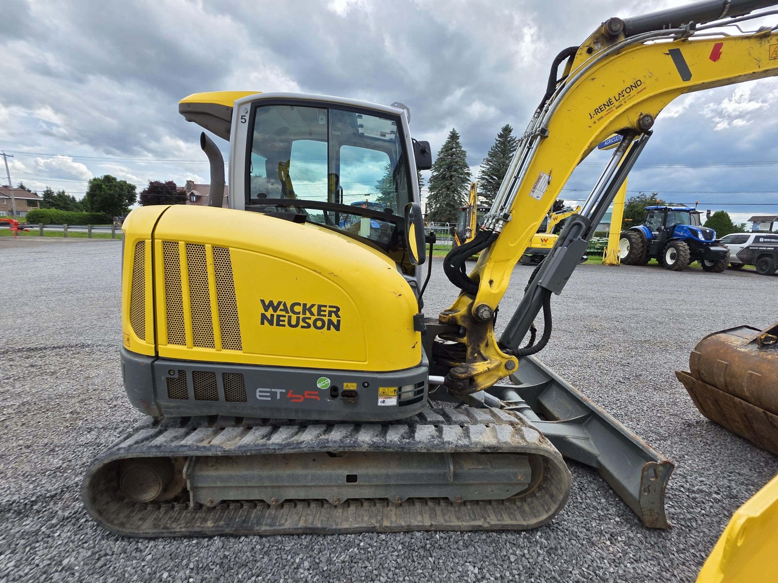 Une pelle compacte Wacker Neuson ET45 jaune est posée sur du gravier, le bras tendu. L'arrière-plan présente des arbres, des bâtiments et d'autres machines sous un ciel nuageux.