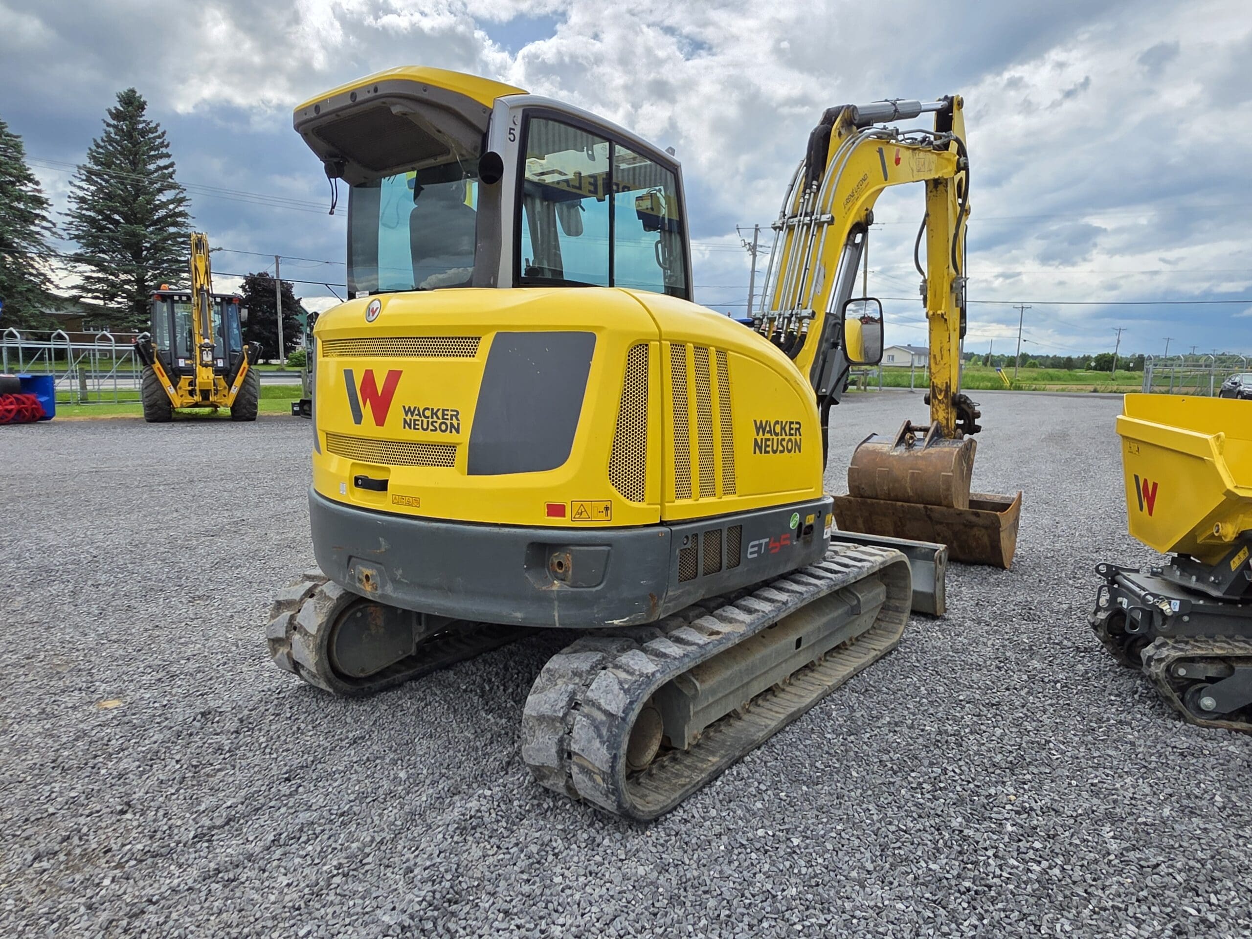 Une pelleteuse Wacker Neuson jaune avec des chenilles en caoutchouc est garée sur du gravier sous un ciel nuageux, avec de la verdure et d'autres équipements de construction en arrière-plan.