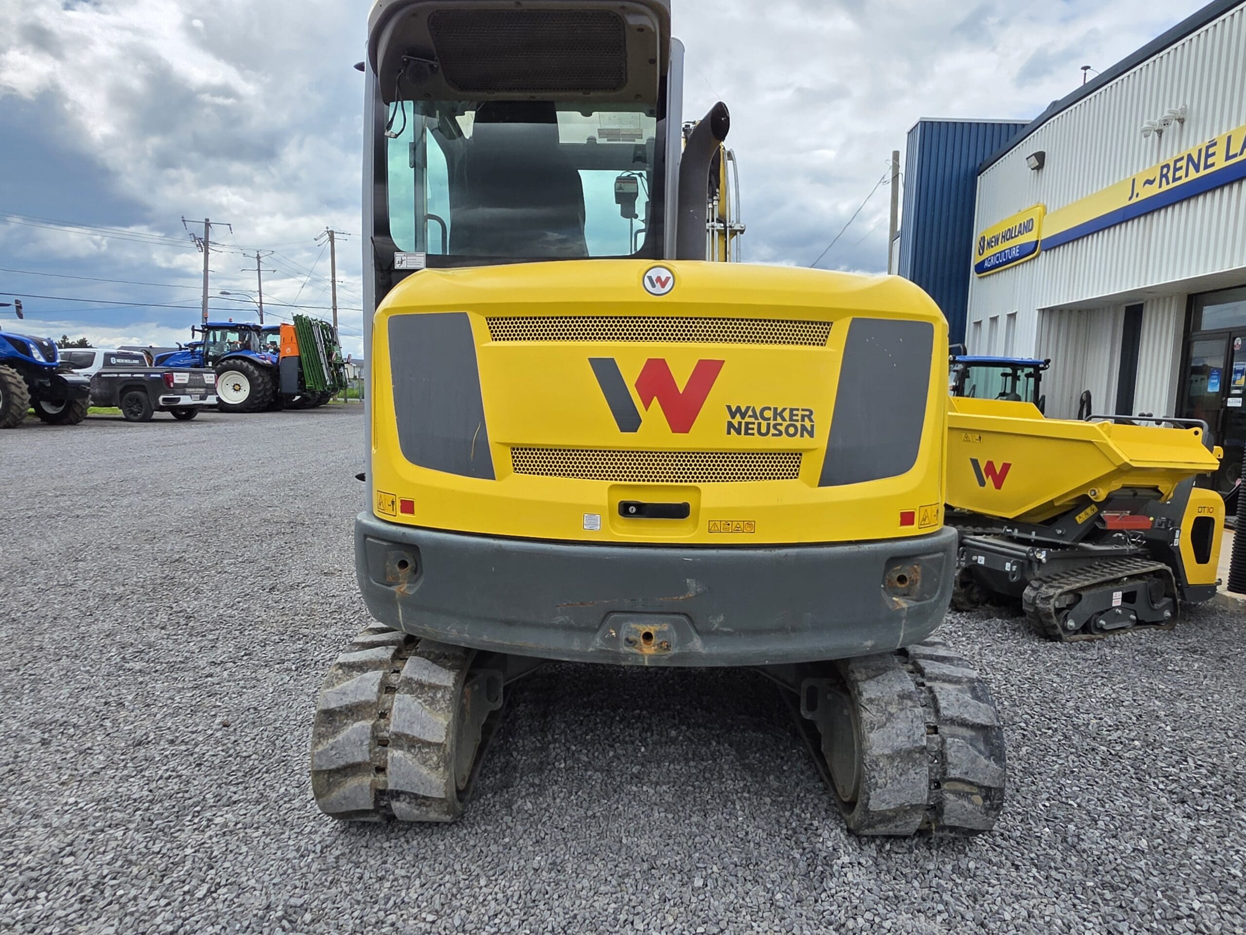 Un véhicule de construction Wacker Neuson jaune est garé sur du gravier à l'extérieur d'un bâtiment, vu de l'arrière. D'autres équipements et un tracteur sont visibles à l'arrière-plan sous un ciel nuageux.