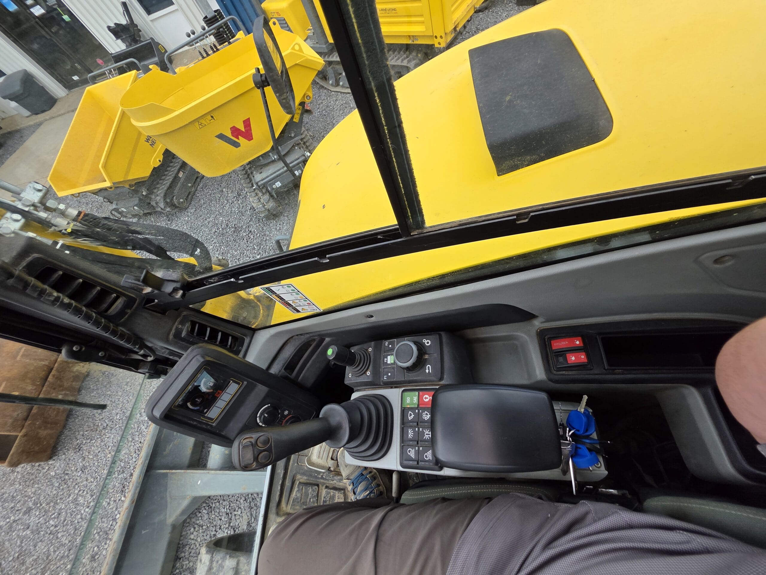Vue de l'intérieur de la cabine d'un véhicule de construction, montrant des panneaux de commande, un joystick et le bras d'une personne. À l'extérieur, un tombereau jaune et des engins de chantier sont visibles sur une surface gravillonnée.