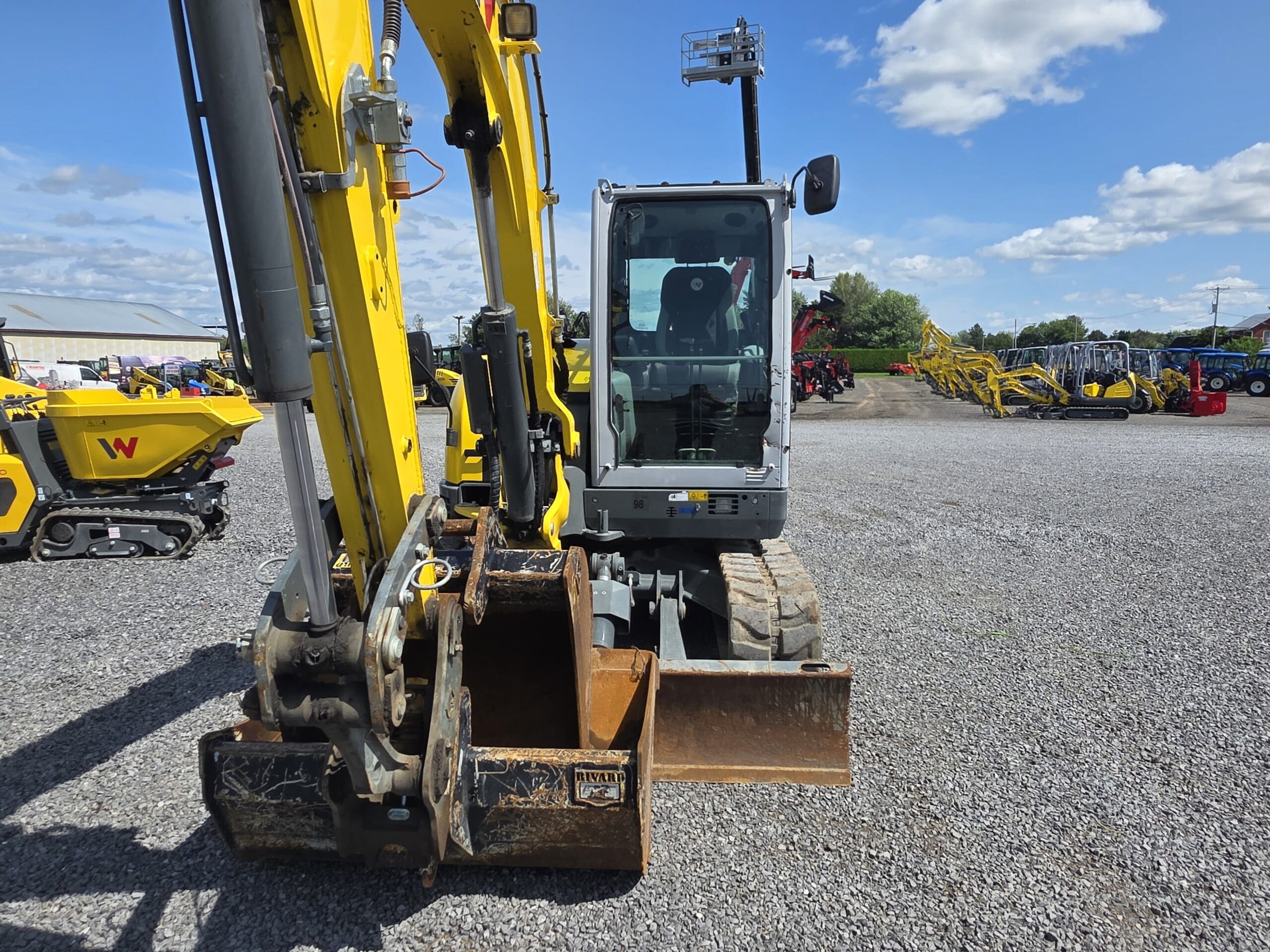Une pelleteuse jaune équipée d'un godet en métal est garée sur un terrain en gravier sous un ciel bleu avec des nuages épars. D'autres machines lourdes sont visibles à l'arrière-plan.