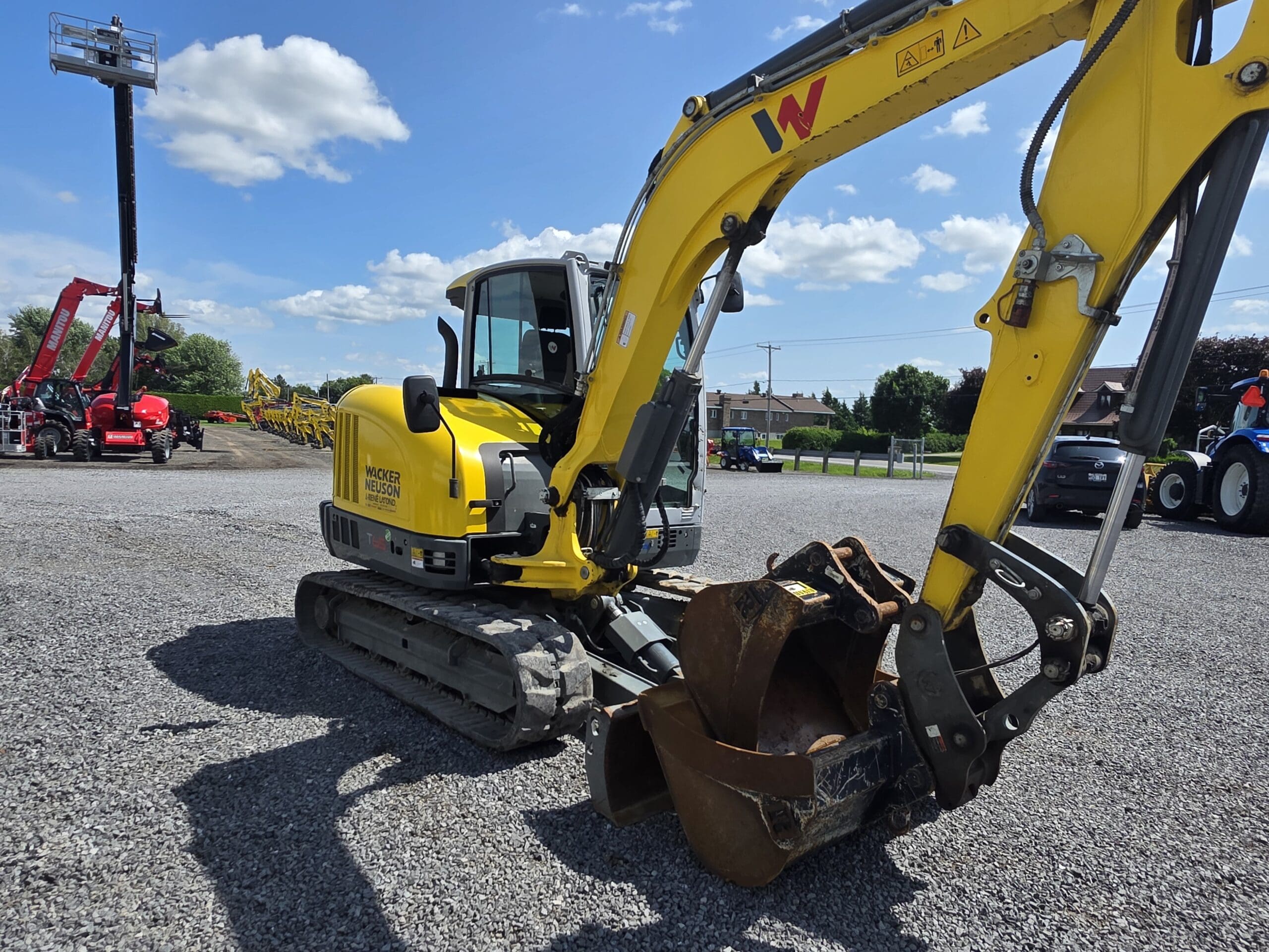 Une pelleteuse compacte jaune avec un godet en métal est garée sur un terrain en gravier sous un ciel bleu avec des nuages épars. D'autres engins de chantier sont visibles à l'arrière-plan.