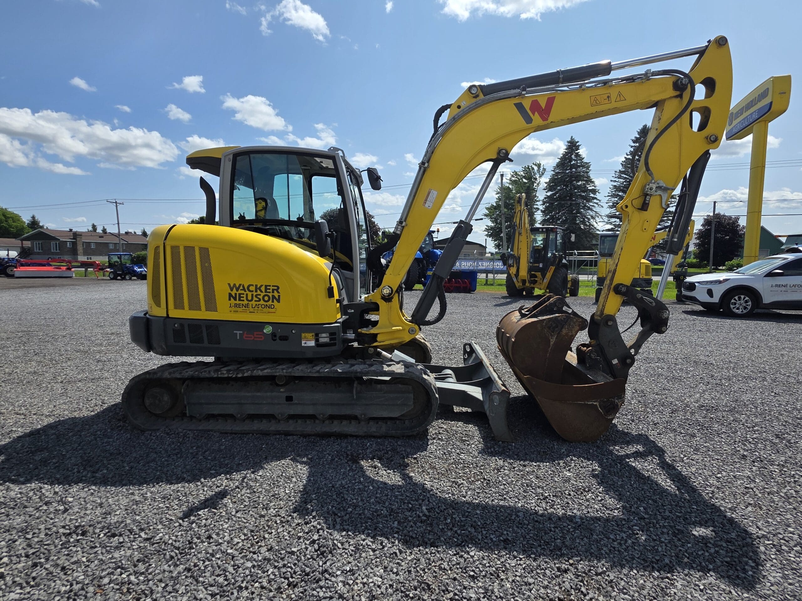 Une mini-pelle Wacker Neuson jaune est garée sur du gravier, son godet reposant sur le sol. Des arbres, des voitures et des lignes électriques sont visibles à l'arrière-plan sous un ciel bleu avec des nuages.