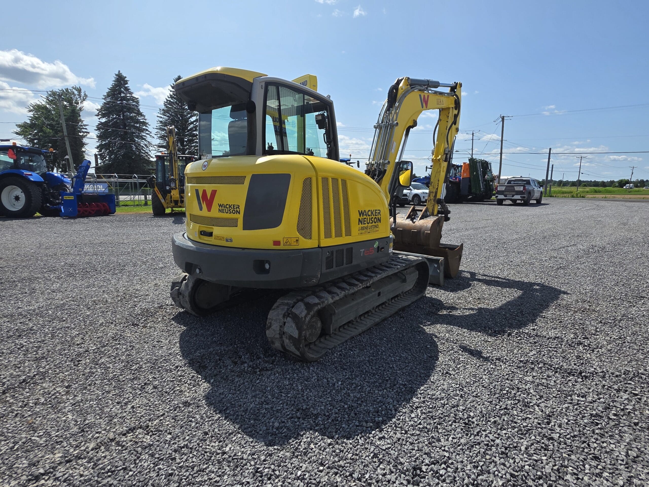 Une mini-pelle Wacker Neuson jaune est garée sur un terrain en gravier sous un ciel bleu clair, avec d'autres machines et véhicules visibles à l'arrière-plan.