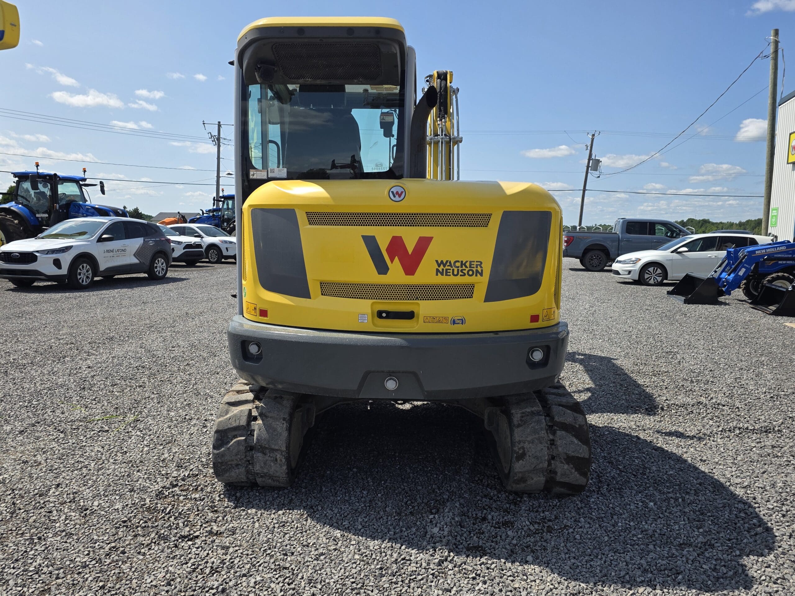 Une pelleteuse Wacker Neuson jaune est garée sur du gravier, photographiée de l'arrière. Plusieurs voitures et tracteurs en stationnement sont visibles à l'arrière-plan sous un ciel partiellement nuageux.