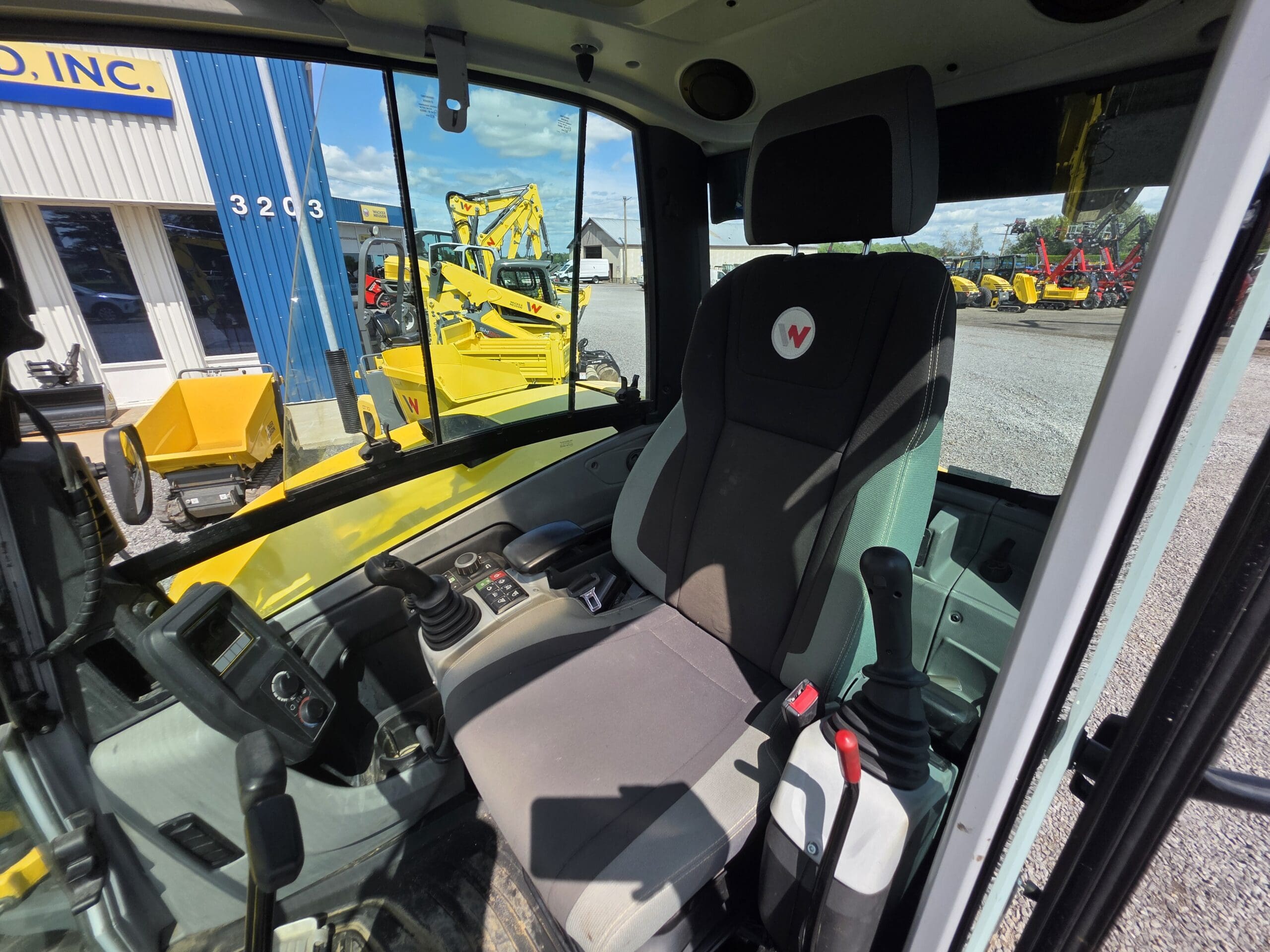 Vue de l'intérieur de la cabine d'un véhicule de construction, montrant un grand siège de conducteur, des manettes de commande et divers boutons. Des engins de chantier jaunes sont visibles à l'extérieur par les fenêtres.