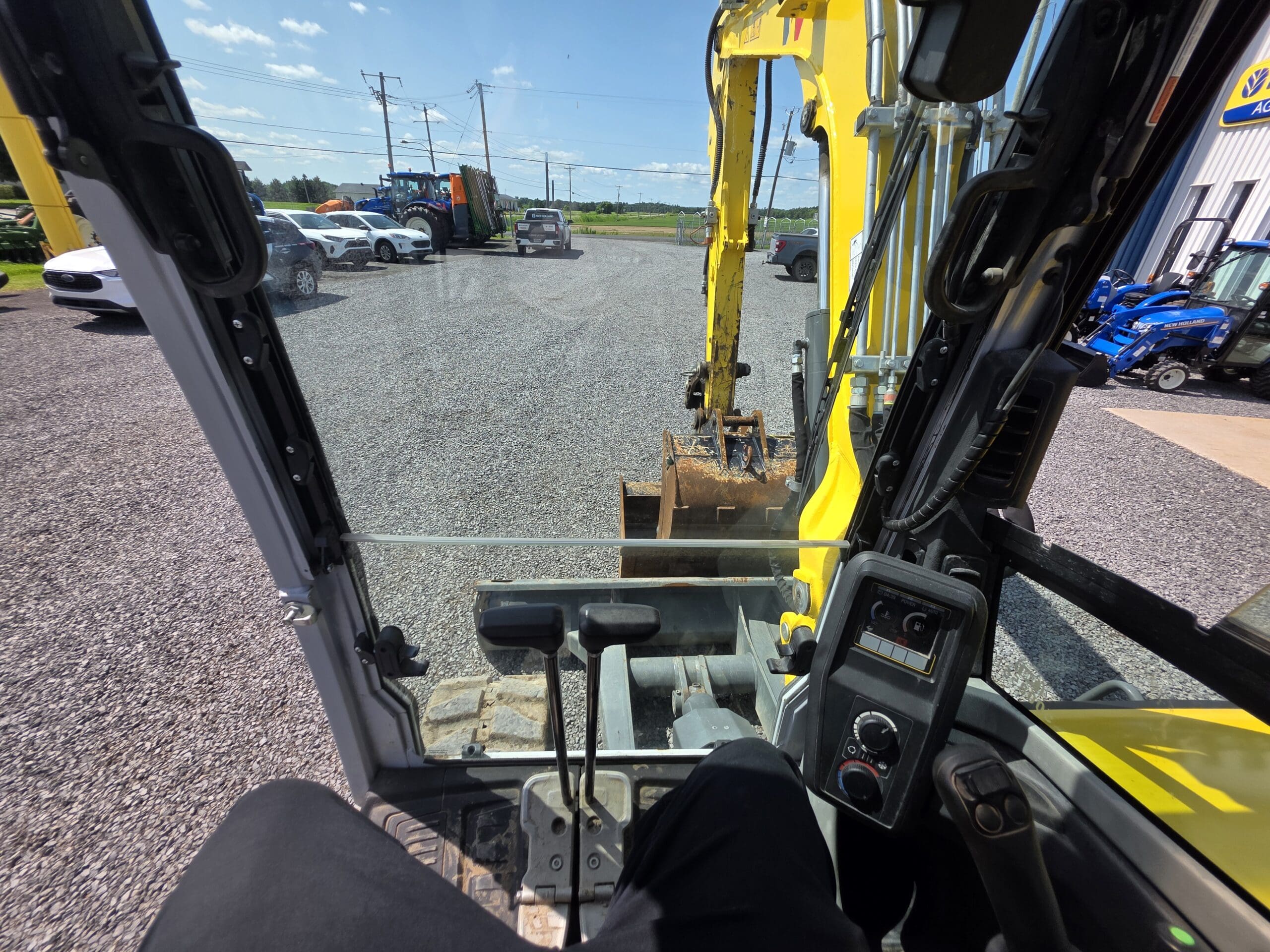 Vue de l'intérieur de la cabine d'une pelleteuse jaune, montrant les commandes, les pédales et le bras de creusement positionné au-dessus d'un terrain de gravier avec des véhicules et des équipements stationnés à l'extérieur sous un ciel bleu.