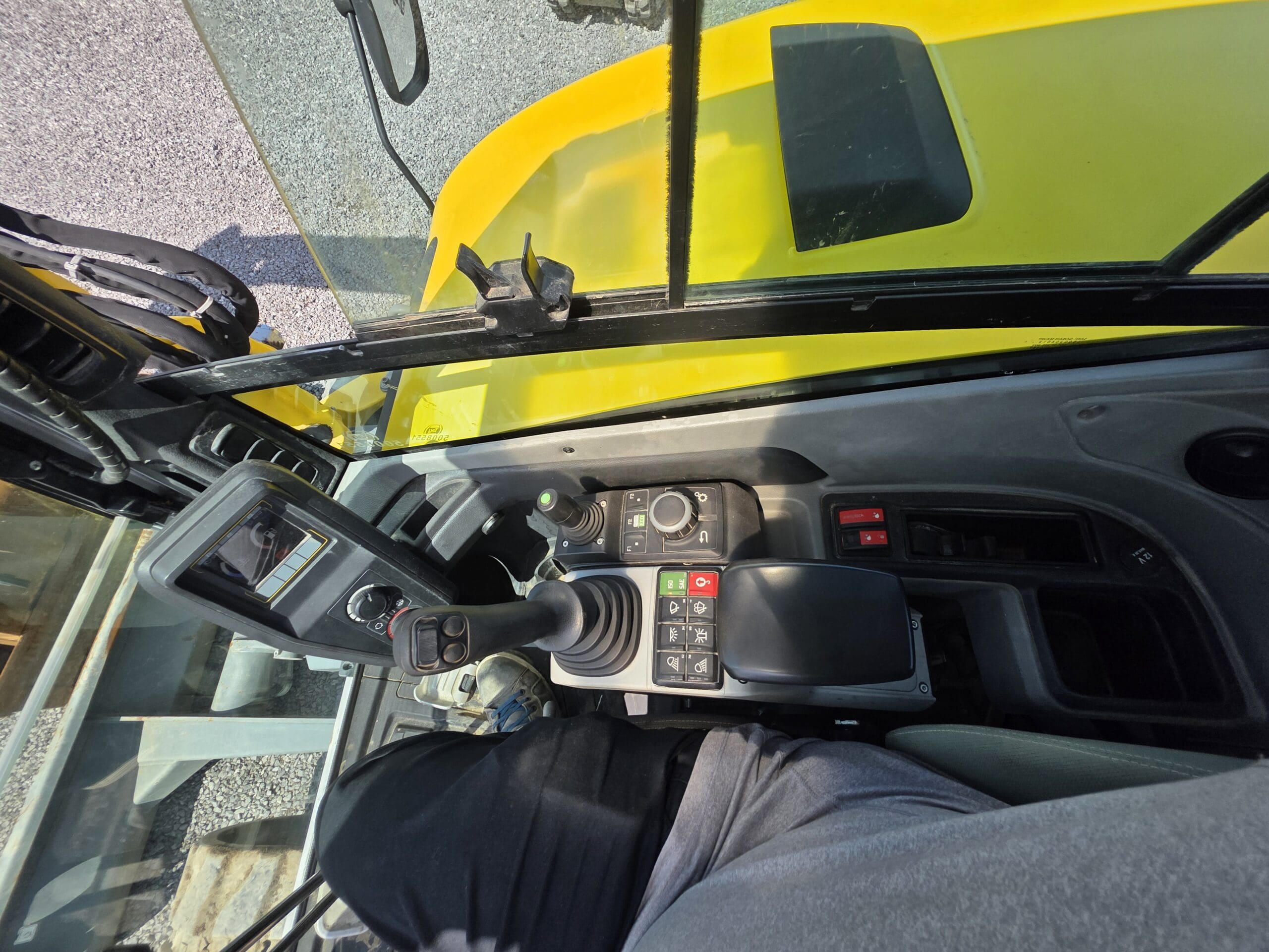 Vue de l'intérieur de la cabine d'un véhicule de chantier jaune, montrant les panneaux de commande, le joystick, les boutons et une partie de la jambe et de la main de l'opérateur. La lumière du soleil éclaire les commandes à travers les fenêtres.