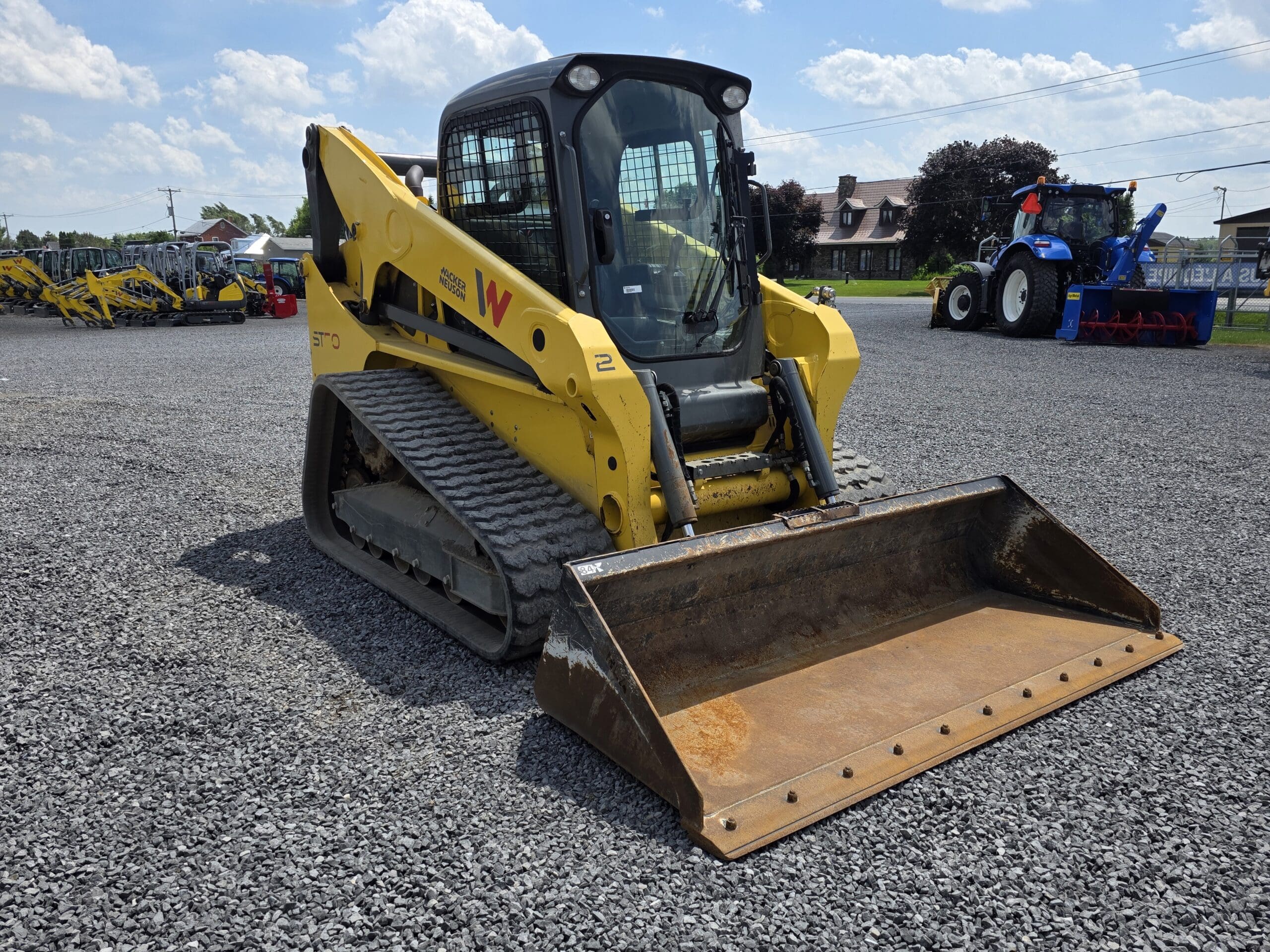 Une chargeuse compacte sur chenilles jaune équipée d'un godet avant est garée sur un terrain en gravier. D'autres machines et un tracteur bleu sont visibles à l'arrière-plan sous un ciel partiellement nuageux.
