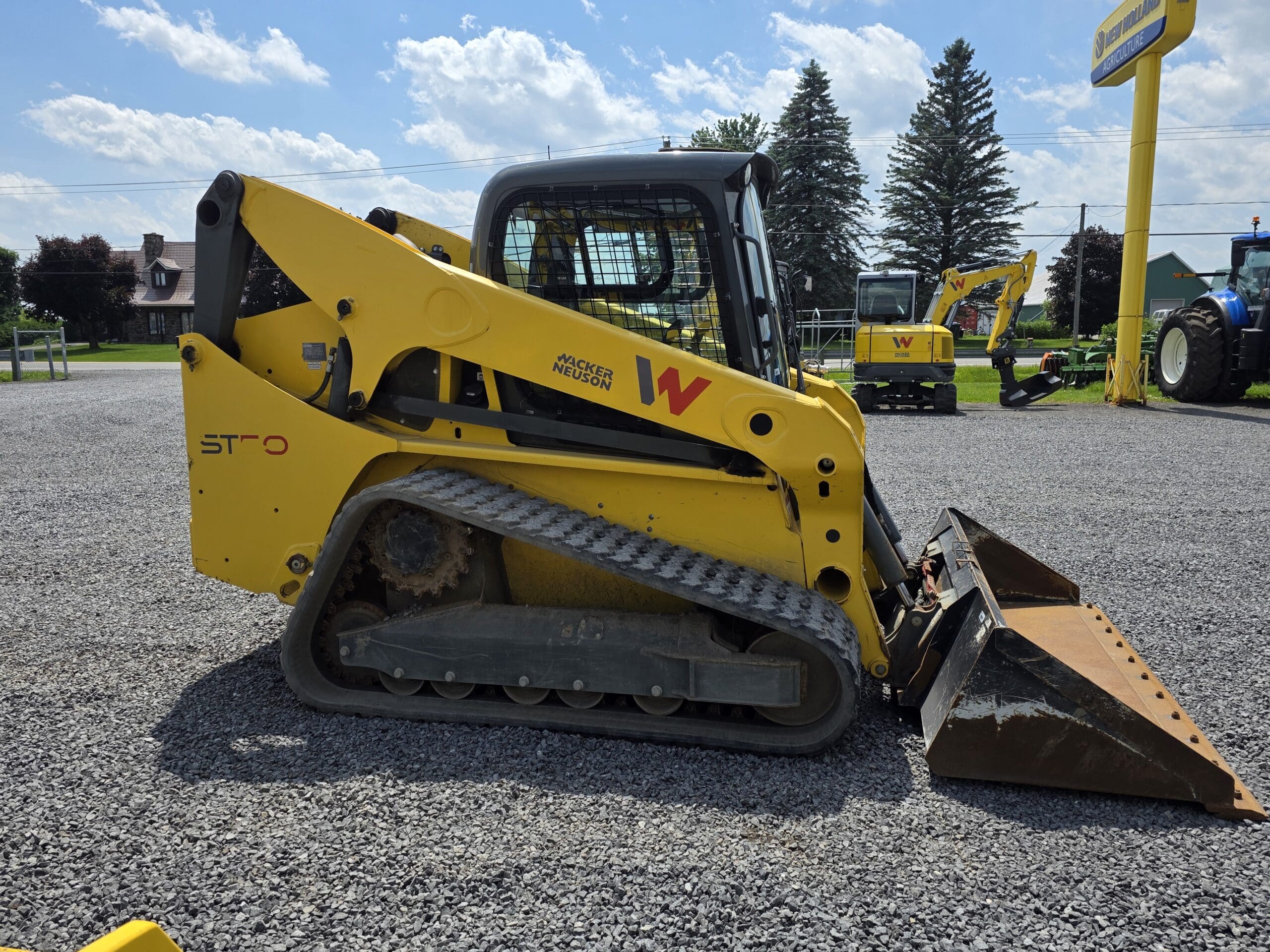 Une chargeuse compacte Wacker Neuson jaune, équipée de chenilles en caoutchouc et d'un godet avant, est garée sur du gravier, avec d'autres machines et des arbres visibles à l'arrière-plan.