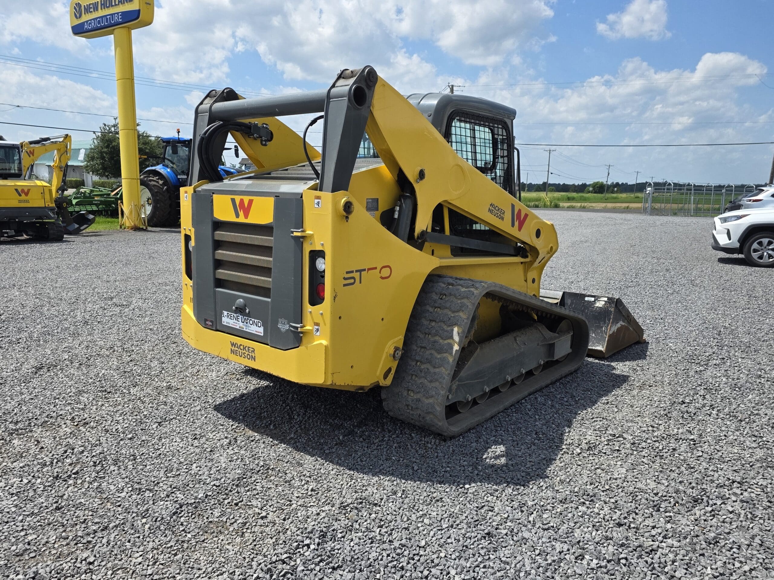 Une chargeuse compacte sur chenilles Wacker Neuson ST31 jaune, équipée d'un godet, est garée sur un terrain en gravier sous un ciel partiellement nuageux. D'autres équipements et véhicules sont visibles à l'arrière-plan.