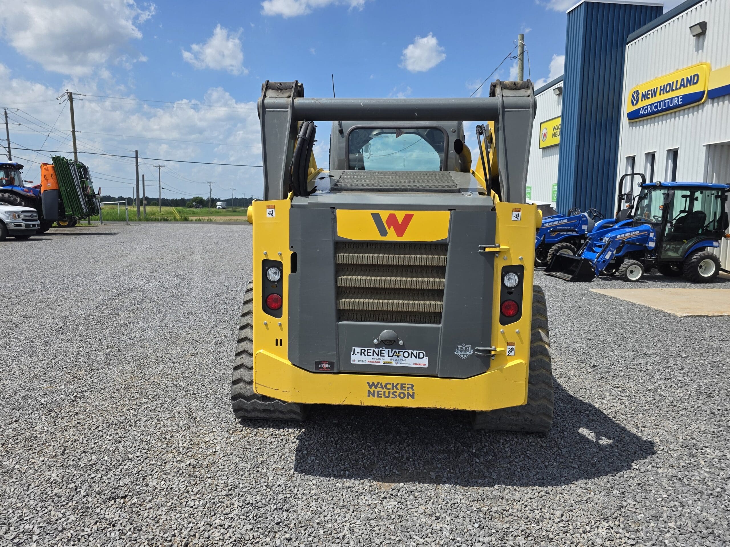 Une chargeuse compacte Wacker Neuson jaune est garée sur du gravier à l'extérieur d'un bâtiment New Holland Agriculture, avec des tracteurs visibles à droite et des lignes électriques en arrière-plan, sous un ciel partiellement nuageux.