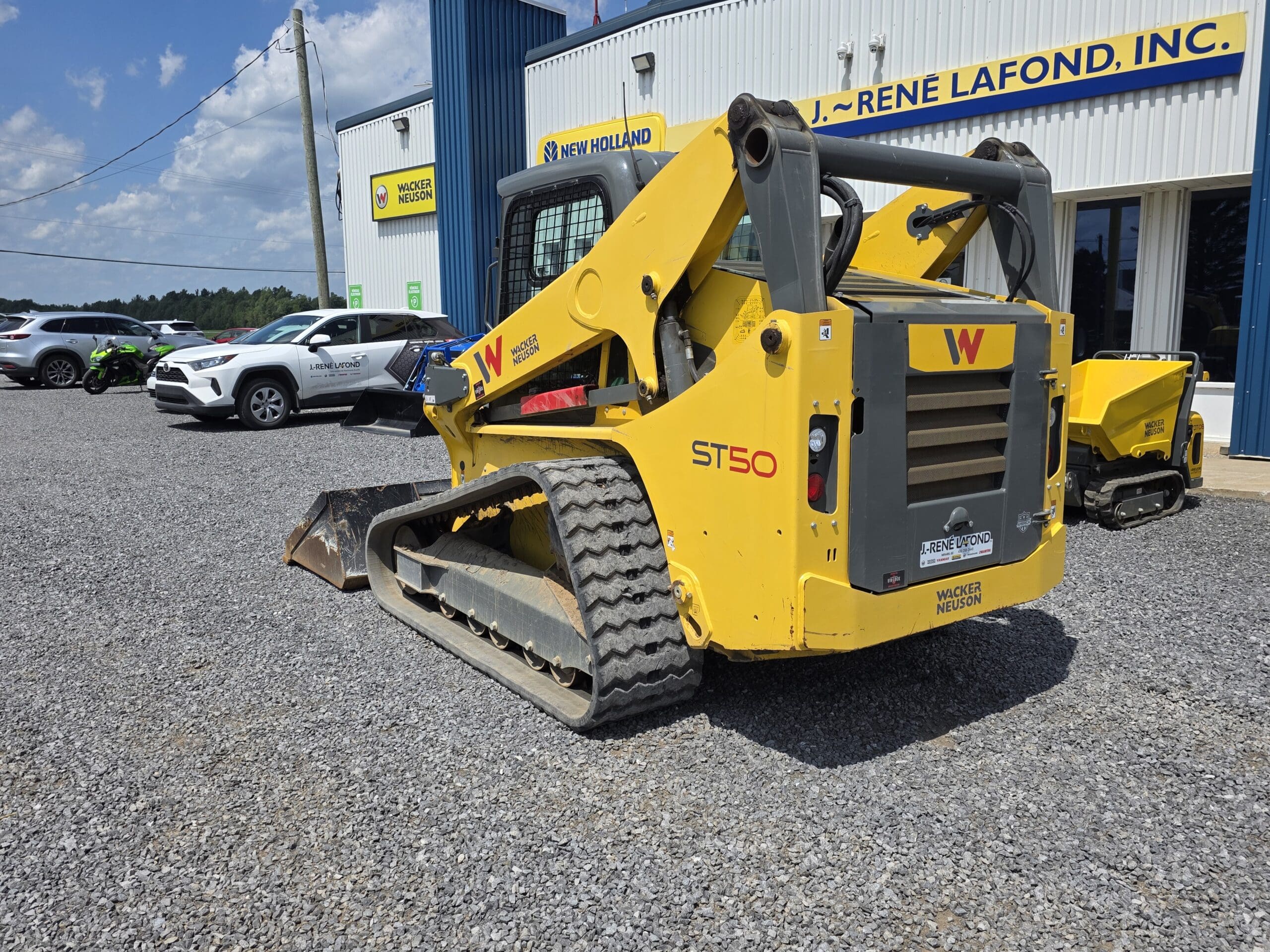 Une chargeuse compacte sur chenilles Wacker Neuson ST50 jaune est garée sur du gravier à l'extérieur d'une concession New Holland par une journée ensoleillée. Plusieurs voitures sont garées à proximité et un bâtiment avec des panneaux de signalisation se trouve à l'arrière-plan.