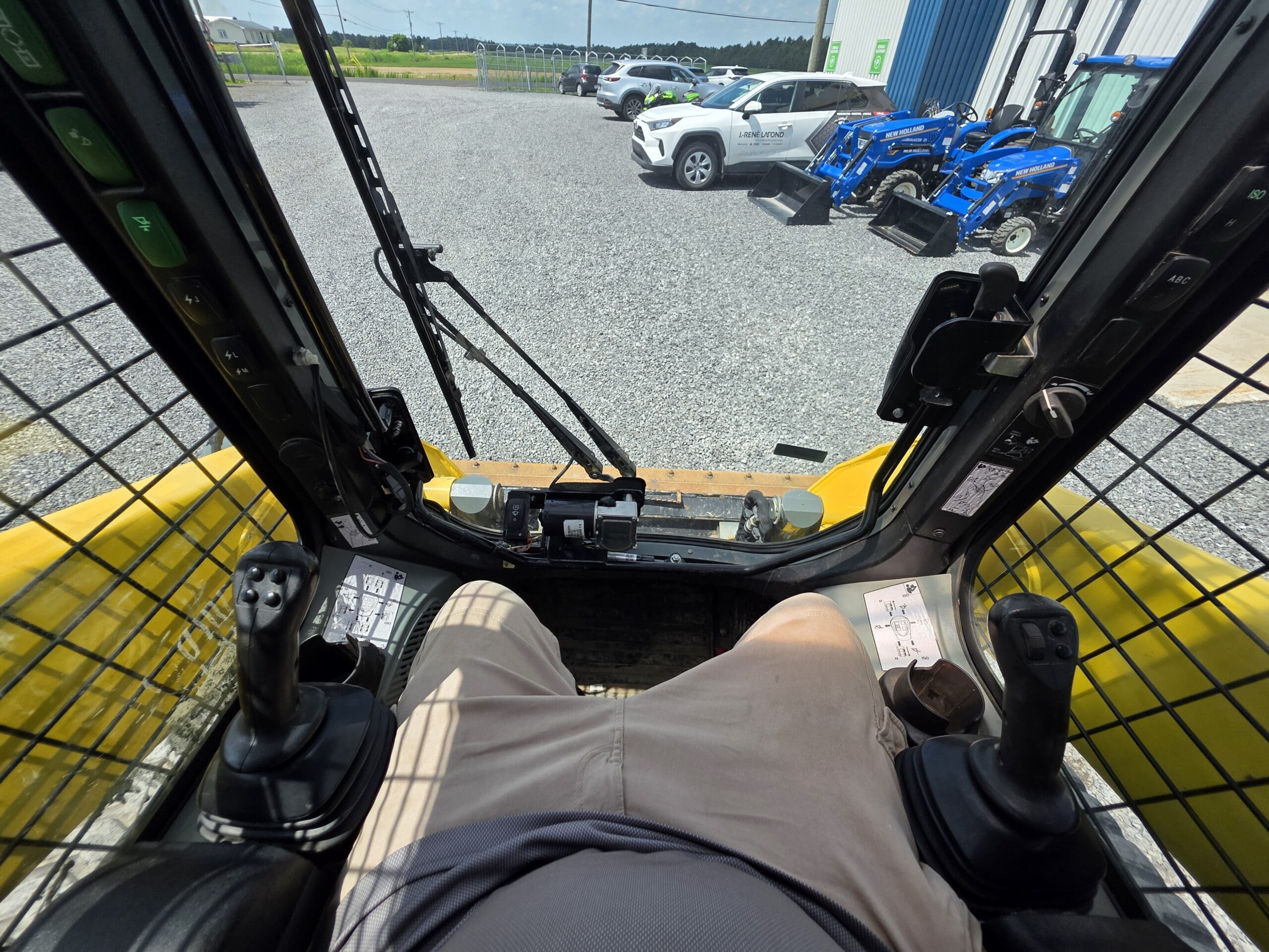 Vue de l'intérieur de la cabine d'un véhicule de chantier, montrant les jambes de l'opérateur, les manettes de commande et le pare-brise. À l'extérieur, on voit des véhicules garés et des machines bleues sur un terrain en gravier.