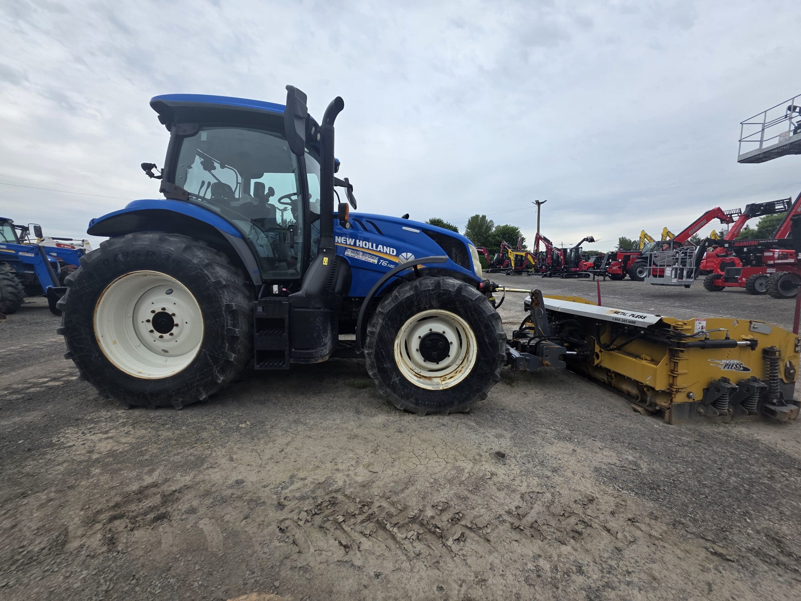 Un tracteur New Holland bleu avec de gros pneus noirs est garé sur un terrain en terre battue, attaché à une machine jaune. D'autres tracteurs et équipements sont visibles à l'arrière-plan sous un ciel nuageux.