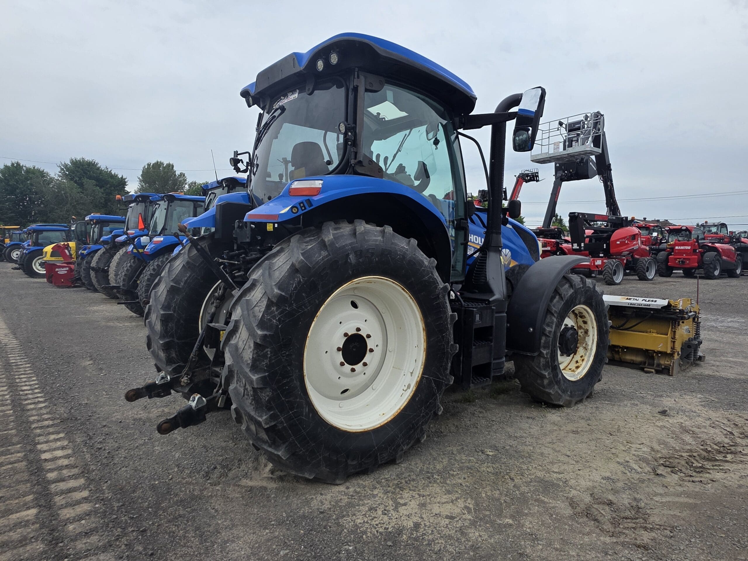 Un grand tracteur bleu est garé sur un terrain en gravier parmi d'autres véhicules et machines agricoles, sous un ciel nuageux. Le tracteur est vu de l'arrière, montrant ses gros pneus et sa cabine.