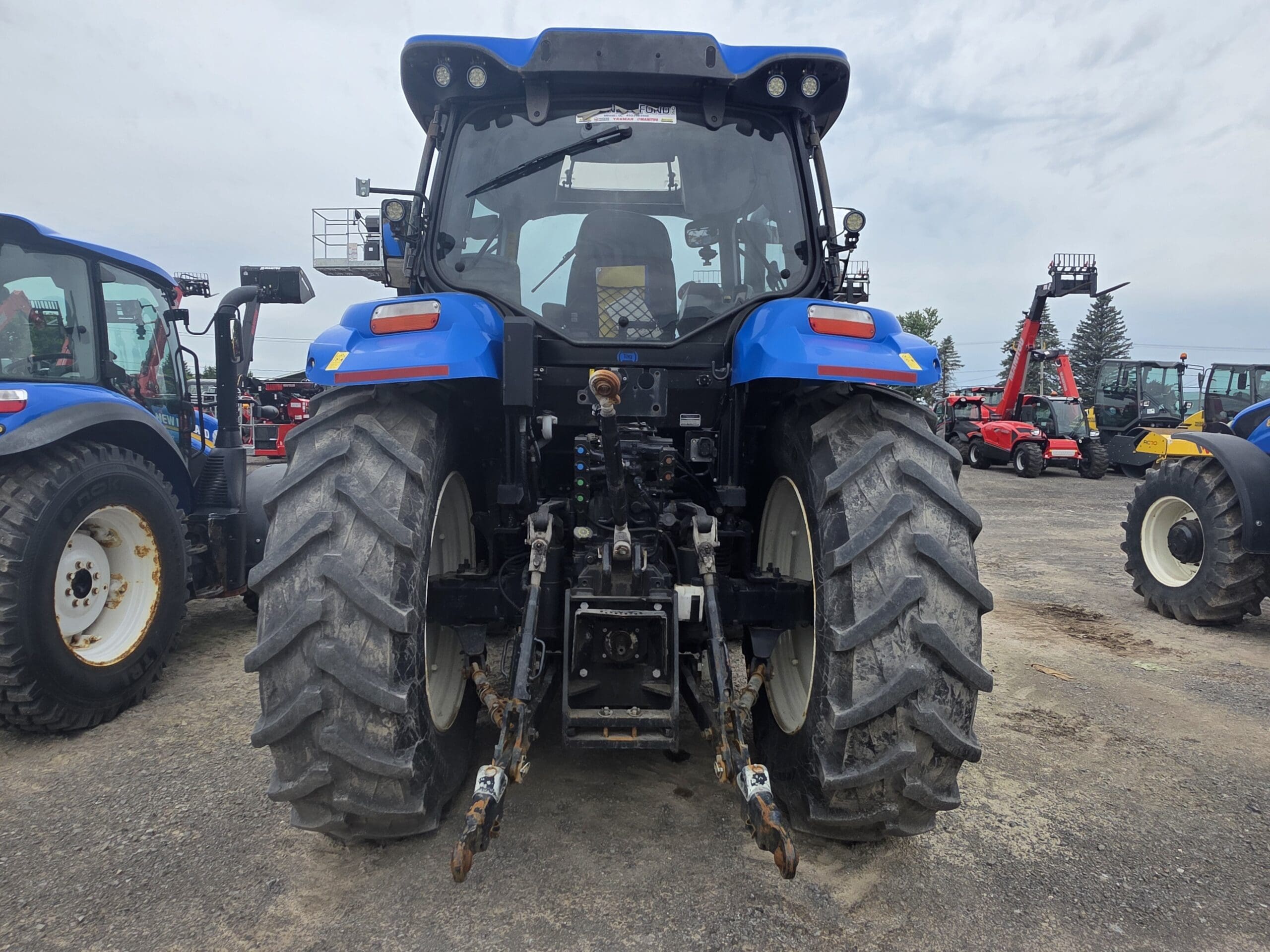 Vue arrière d'un tracteur bleu à gros pneus, garé sur un terrain en gravier parmi d'autres équipements et véhicules agricoles, sous un ciel nuageux.