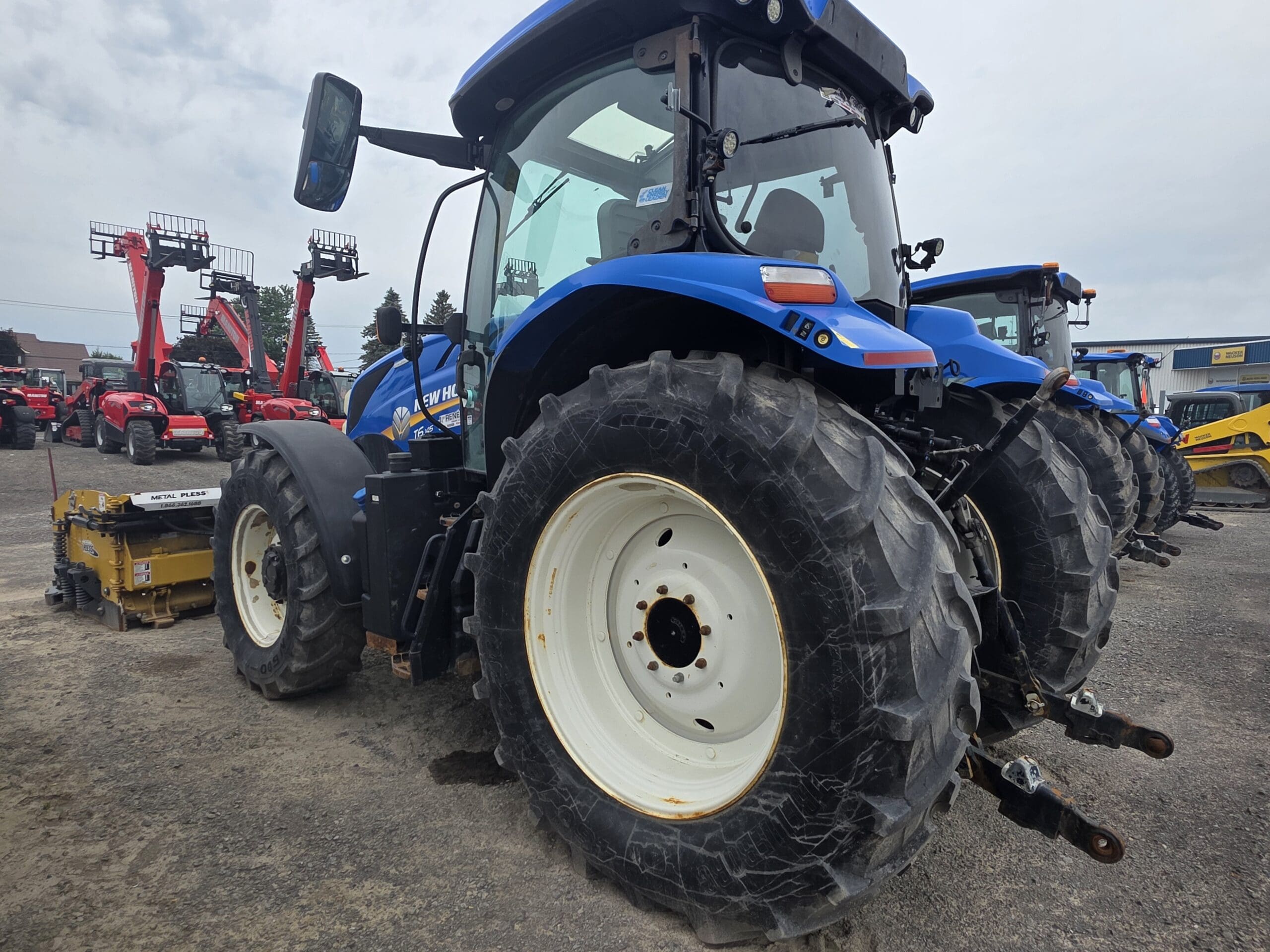 Un gros tracteur bleu aux gros pneus noirs est garé sur un terrain en gravier, entouré d'autres machines et équipements agricoles, sous un ciel nuageux.