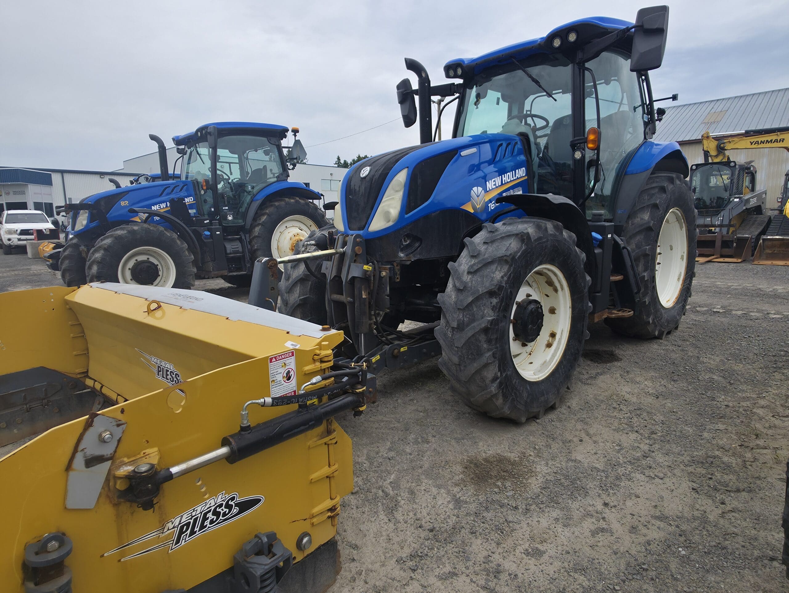 Un tracteur New Holland bleu est garé sur du gravier près d'un outil Pöttinger jaune. Un autre tracteur bleu est visible à l'arrière-plan, ainsi que des bâtiments et des équipements industriels.