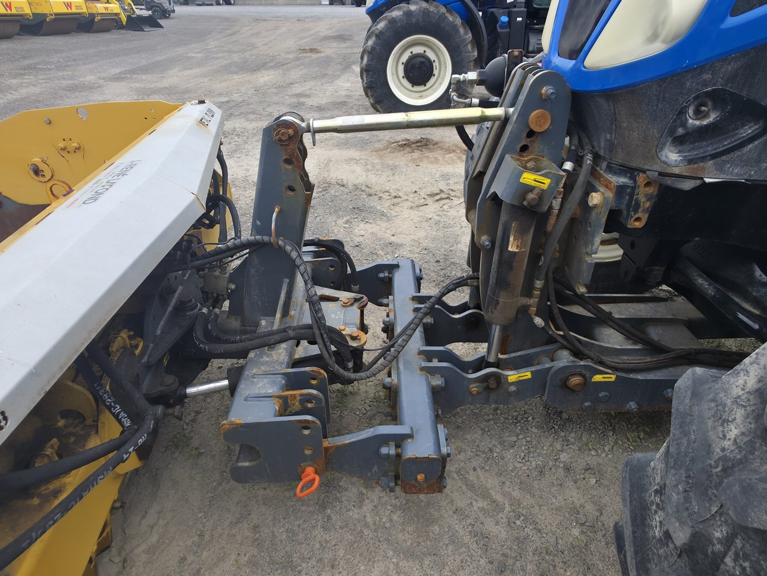 Gros plan du système d'attelage avant d'un tracteur relié à un outil agricole jaune, montrant des tuyaux hydrauliques, des connecteurs métalliques et un peu de rouille sur la machine. Un sol en terre battue et d'autres équipements agricoles sont visibles à l'arrière-plan.
