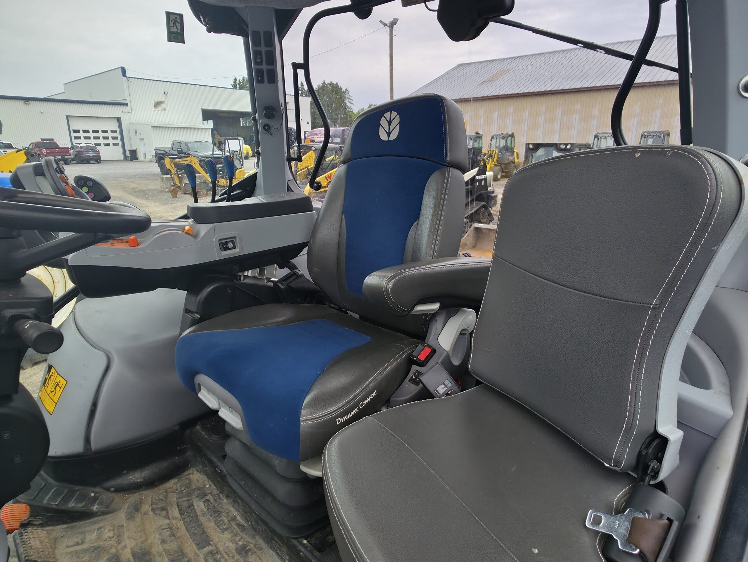L'image montre l'intérieur de la cabine d'un tracteur moderne, avec deux sièges, l'un recouvert de bleu et de noir et l'autre de gris, ainsi que des panneaux de commande, un volant et une vue sur les machines à l'extérieur à travers les fenêtres.