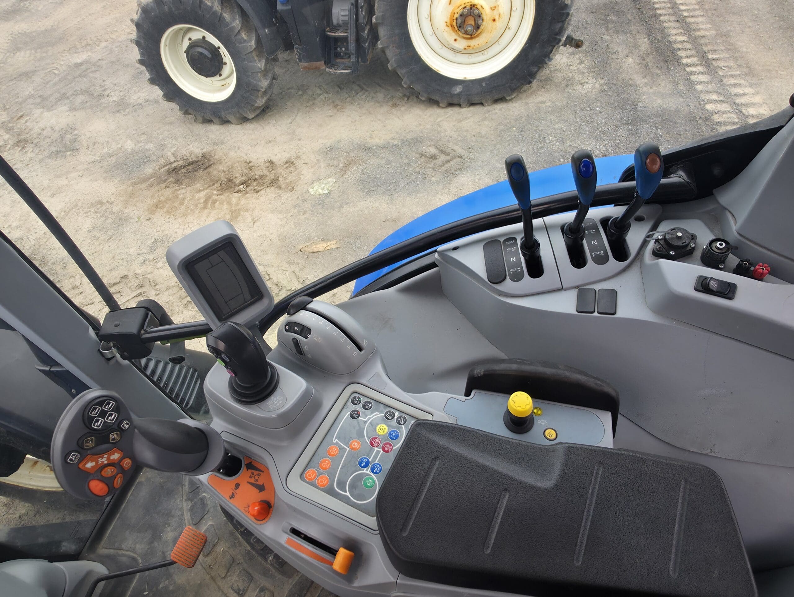 Vue du panneau de commande d'un tracteur montrant divers leviers, boutons, un écran tactile et un joystick à l'intérieur de la cabine. À l'extérieur, une autre roue de tracteur est visible sur une surface de gravier.