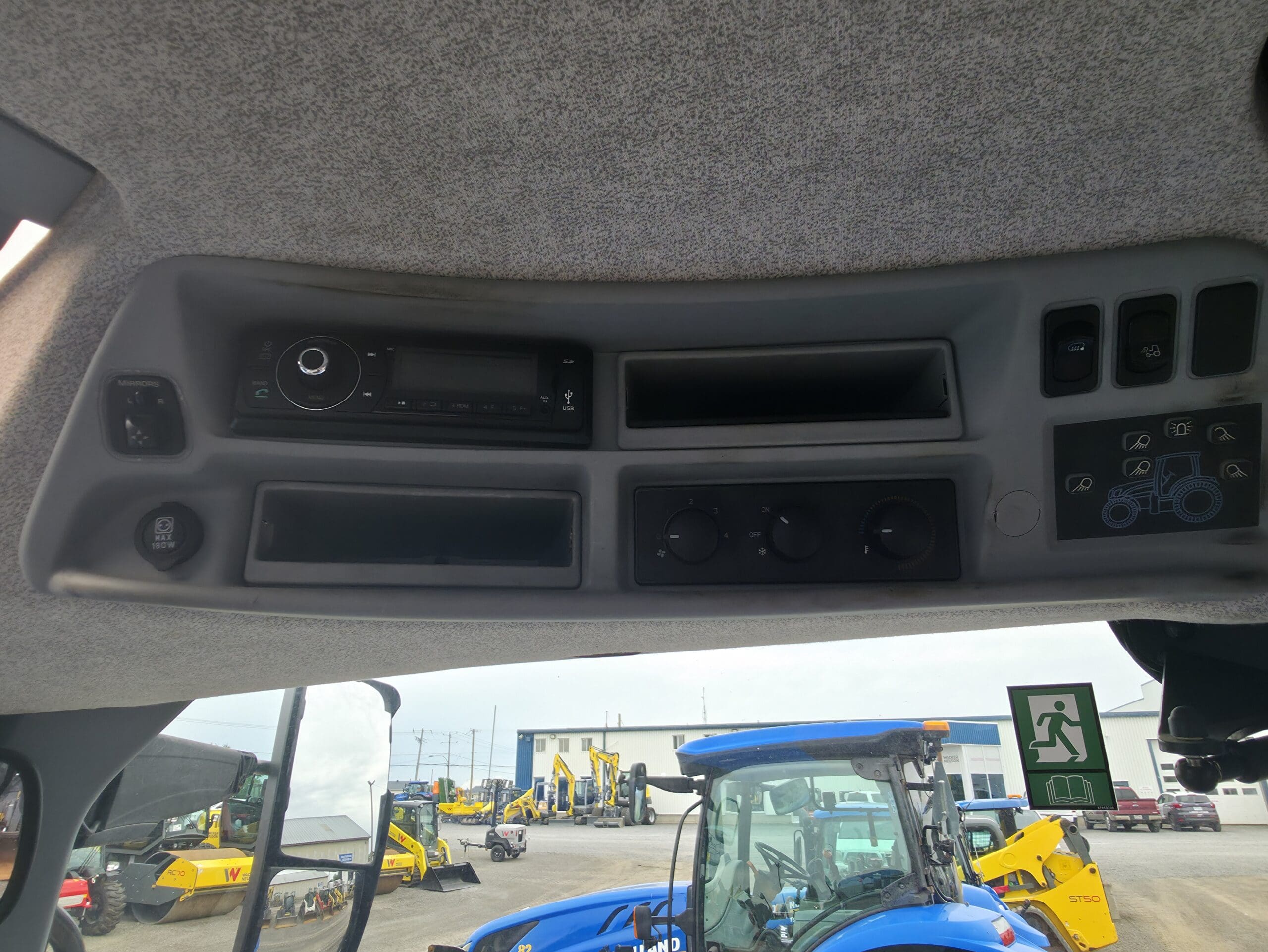 Vue intérieure d'une cabine de tracteur montrant un panneau de commande avec des boutons, des cadrans et des compartiments au-dessus de la tête. Par la fenêtre, on aperçoit à l'extérieur des tracteurs bleus et des engins de chantier jaunes.