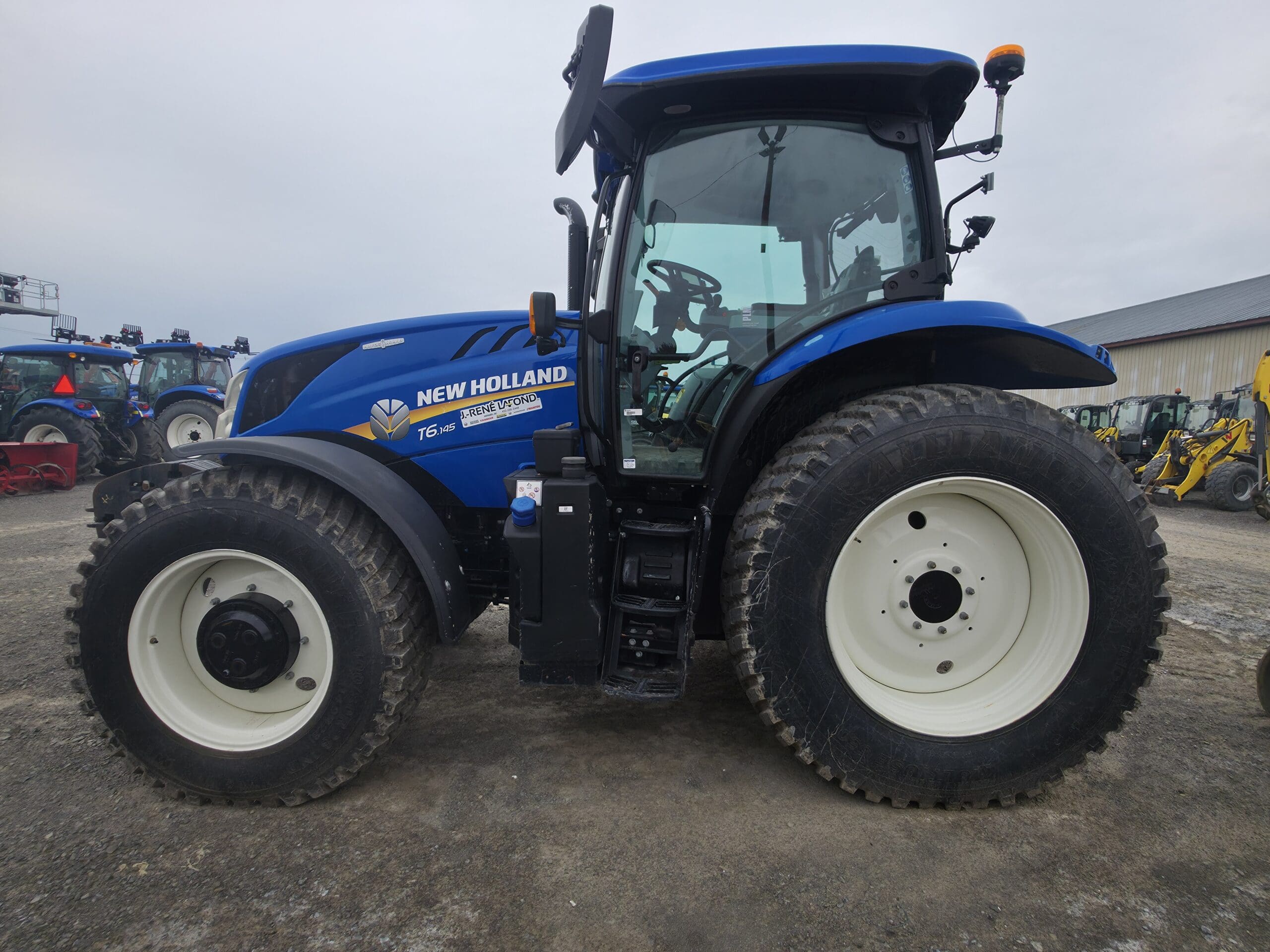 Un tracteur New Holland T6.180 bleu est garé sur un terrain en gravier, en vue latérale. D'autres équipements agricoles et tracteurs sont visibles à l'arrière-plan, près d'un bâtiment.