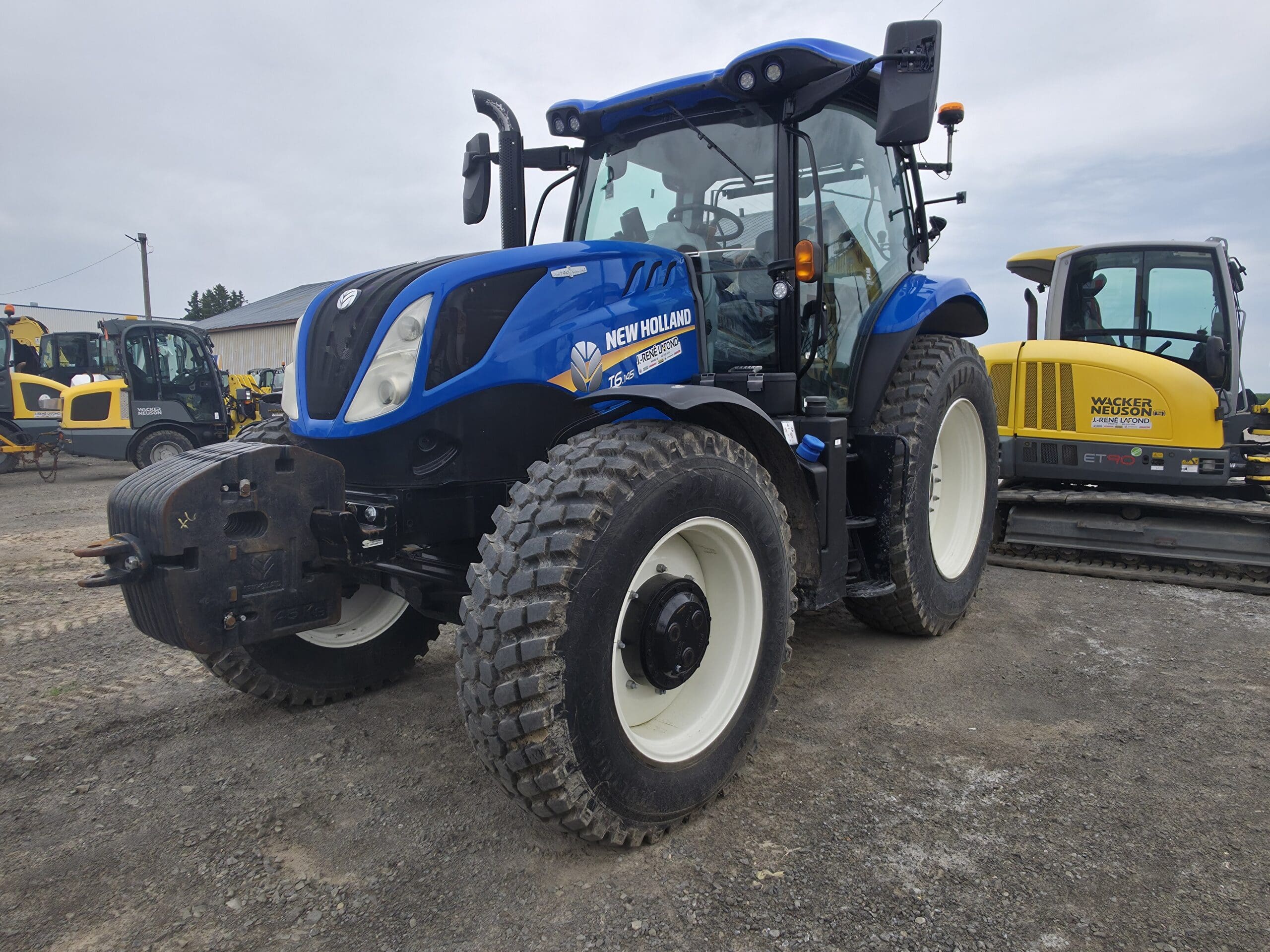 Un tracteur New Holland bleu est garé sur un terrain en gravier, avec du matériel de construction, dont une machine jaune, en arrière-plan, sous un ciel nuageux.