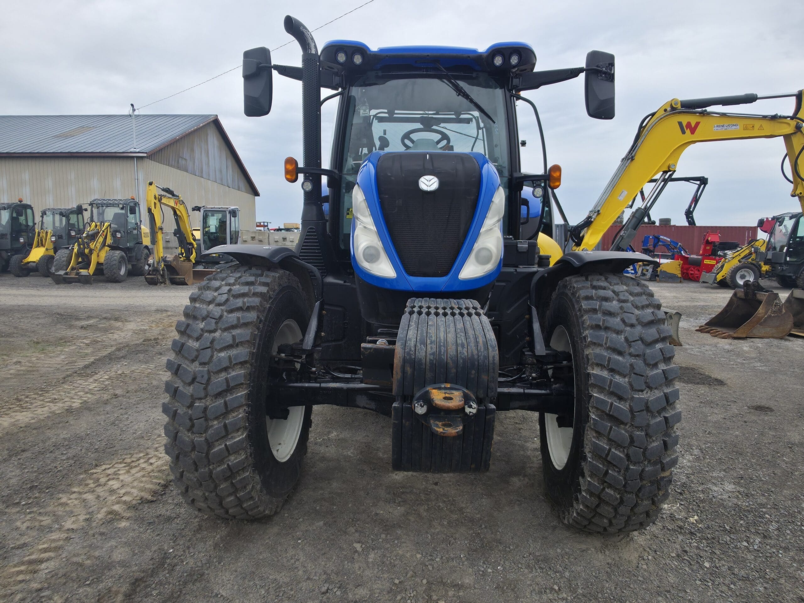 Un grand tracteur bleu à gros pneus est garé sur un terrain en terre battue. Des engins de chantier et un bâtiment métallique sont visibles à l'arrière-plan sous un ciel nuageux.
