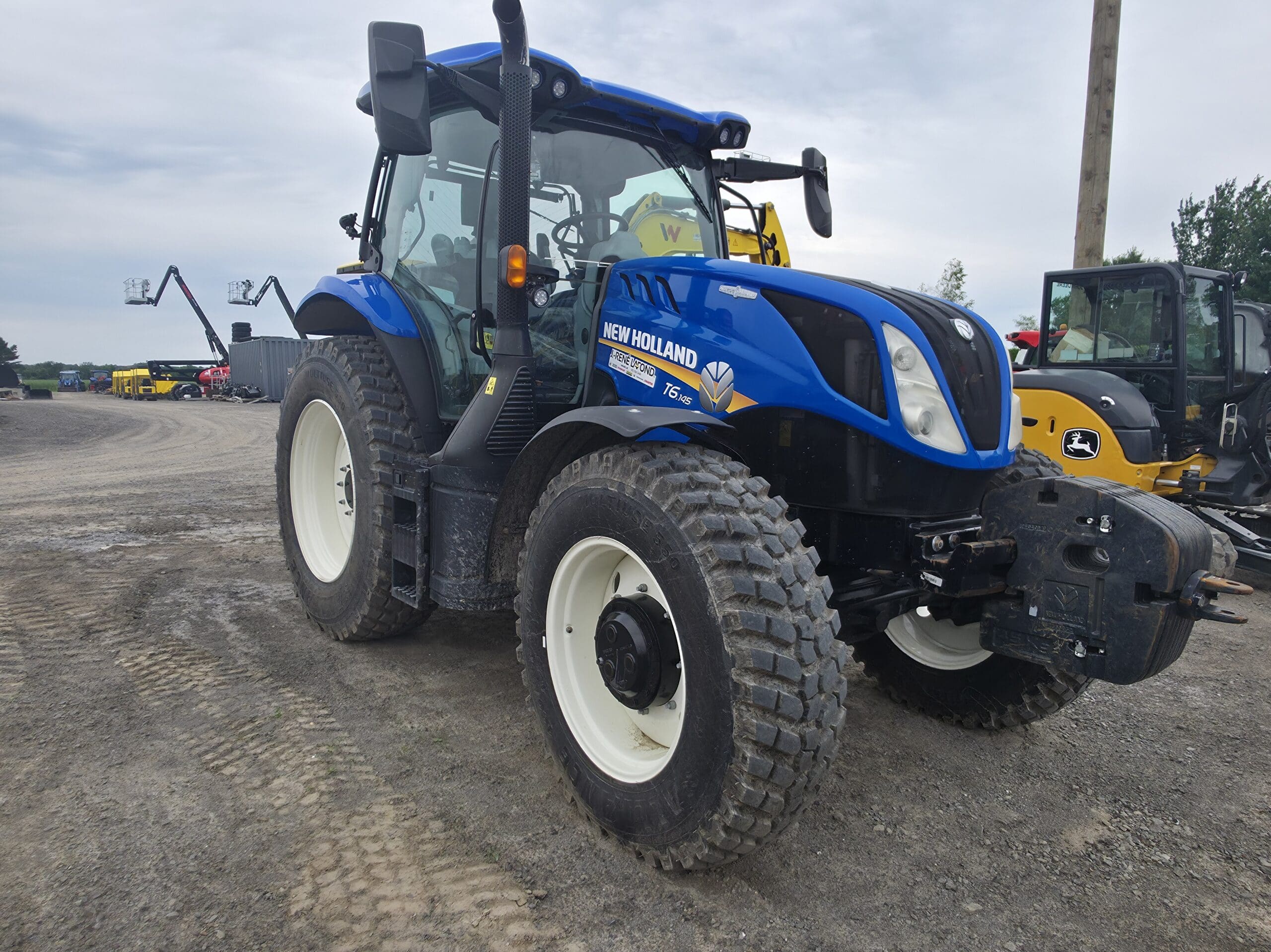 Un tracteur New Holland bleu à gros pneus garé sur un terrain en terre battue, entouré d'engins de chantier et d'équipements lourds, sous un ciel nuageux.