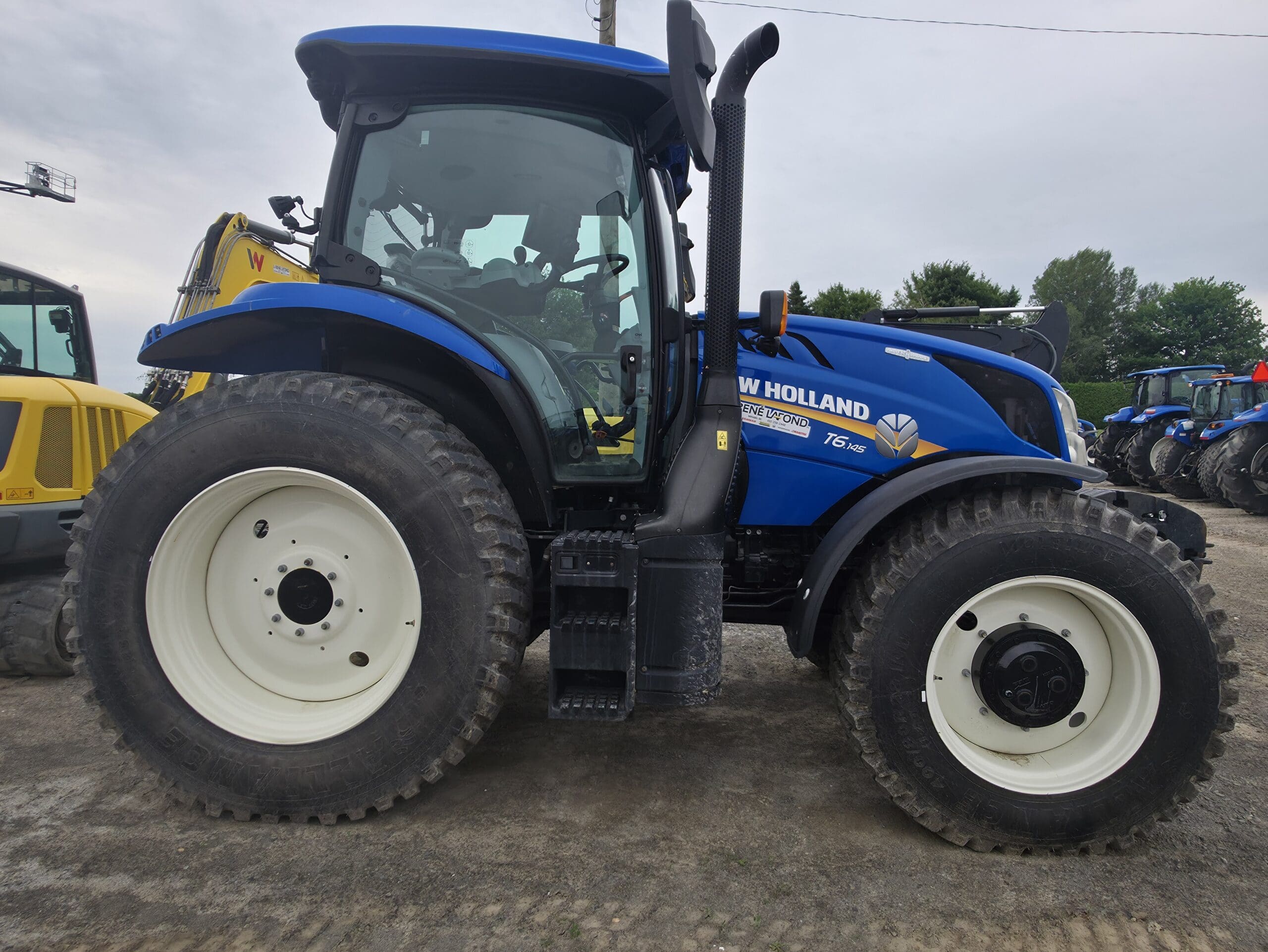 Un tracteur New Holland T6.155 bleu est garé sur un terrain en terre, vu de côté. Le tracteur est équipé de gros pneus robustes et est entouré d'autres machines et d'arbres à l'arrière-plan.