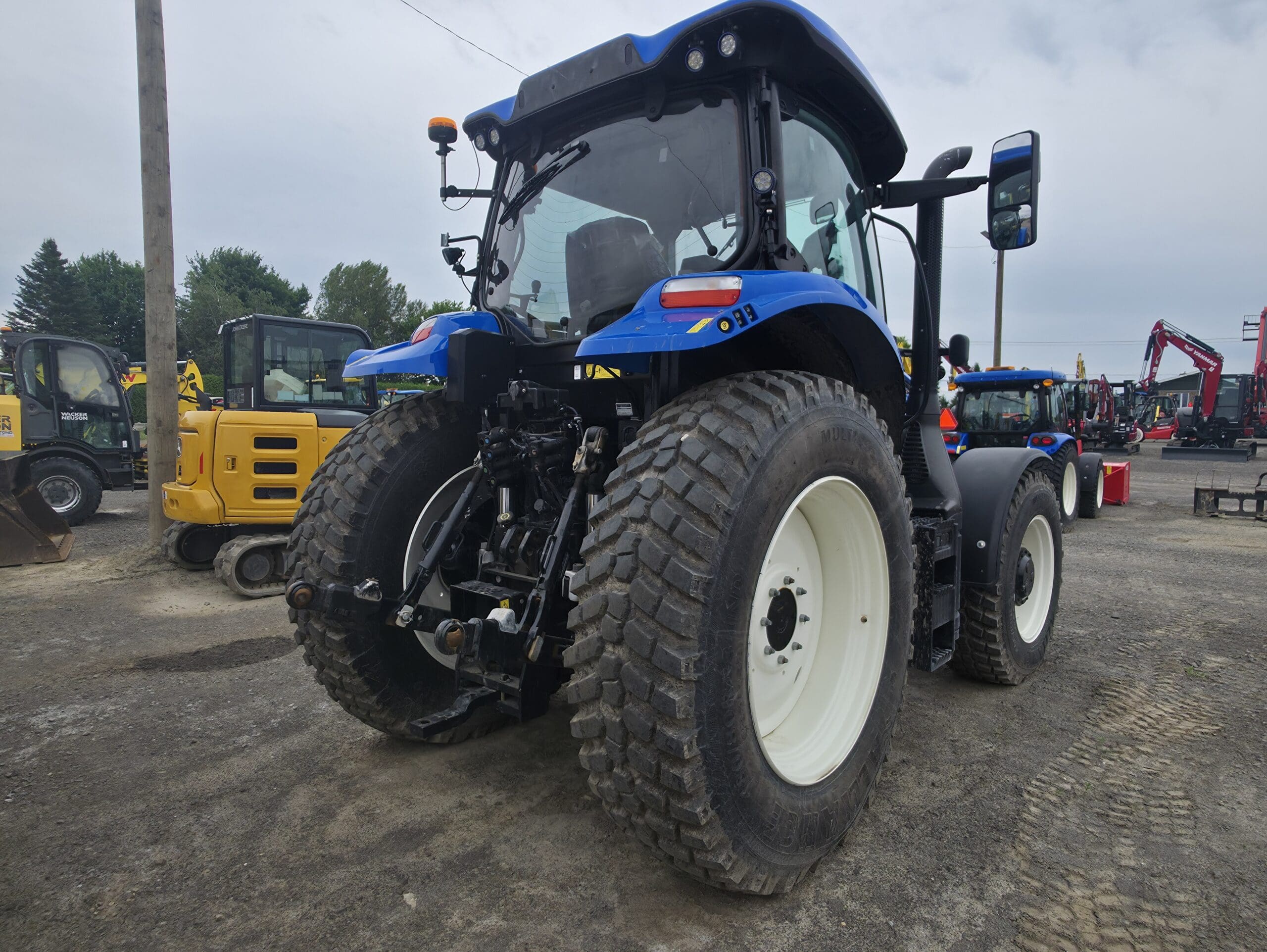 Un tracteur bleu équipé de gros pneus arrière est garé sur un terrain en terre battue à proximité d'autres engins de chantier, dont un véhicule jaune. Des arbres et des machines sont visibles à l'arrière-plan sous un ciel nuageux.