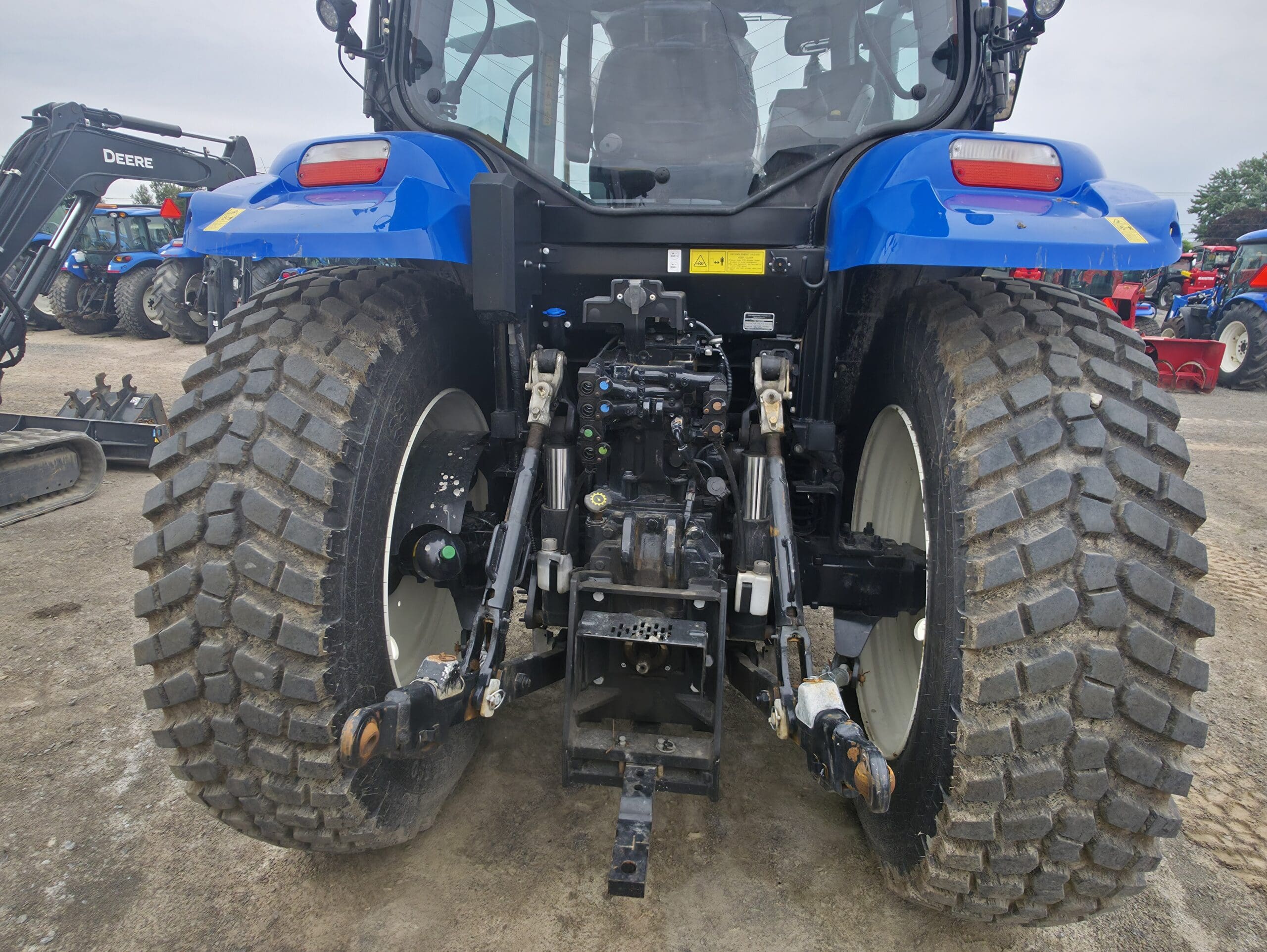 Gros plan sur l'arrière d'un tracteur bleu, montrant de gros pneus texturés et le mécanisme d'attelage. D'autres tracteurs et équipements sont visibles à l'arrière-plan sur un terrain en terre.