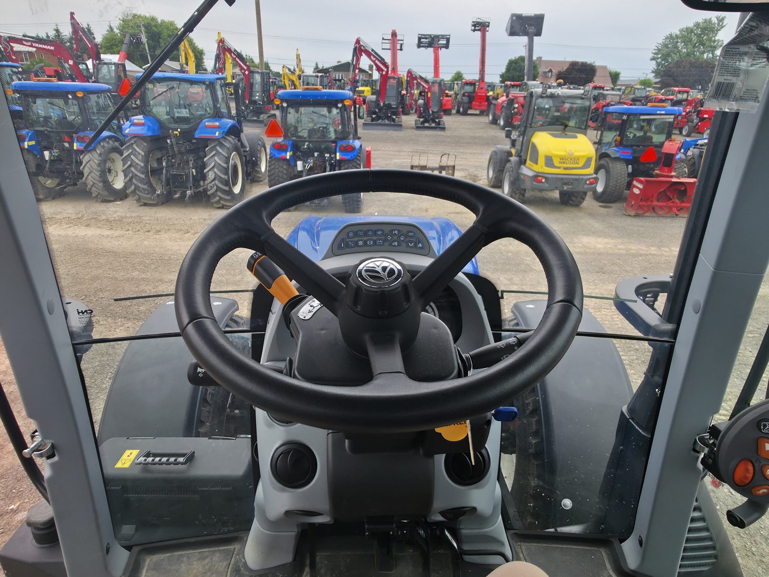 Vue de l'intérieur d'une cabine de tracteur, en regardant par-dessus le volant vers un terrain rempli de tracteurs et de machines lourdes exposés à l'extérieur. Diverses couleurs et modèles d'équipement sont visibles à l'arrière-plan.