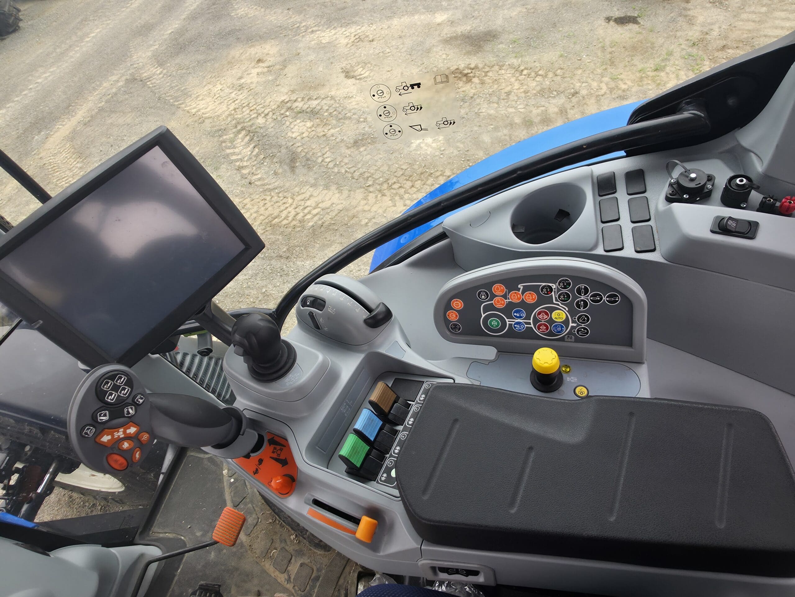 Gros plan du panneau de commande d'un tracteur moderne montrant un écran tactile, un joystick, des boutons de différentes couleurs, des interrupteurs et des leviers à l'intérieur de la cabine. Le sol extérieur est visible à travers la fenêtre.