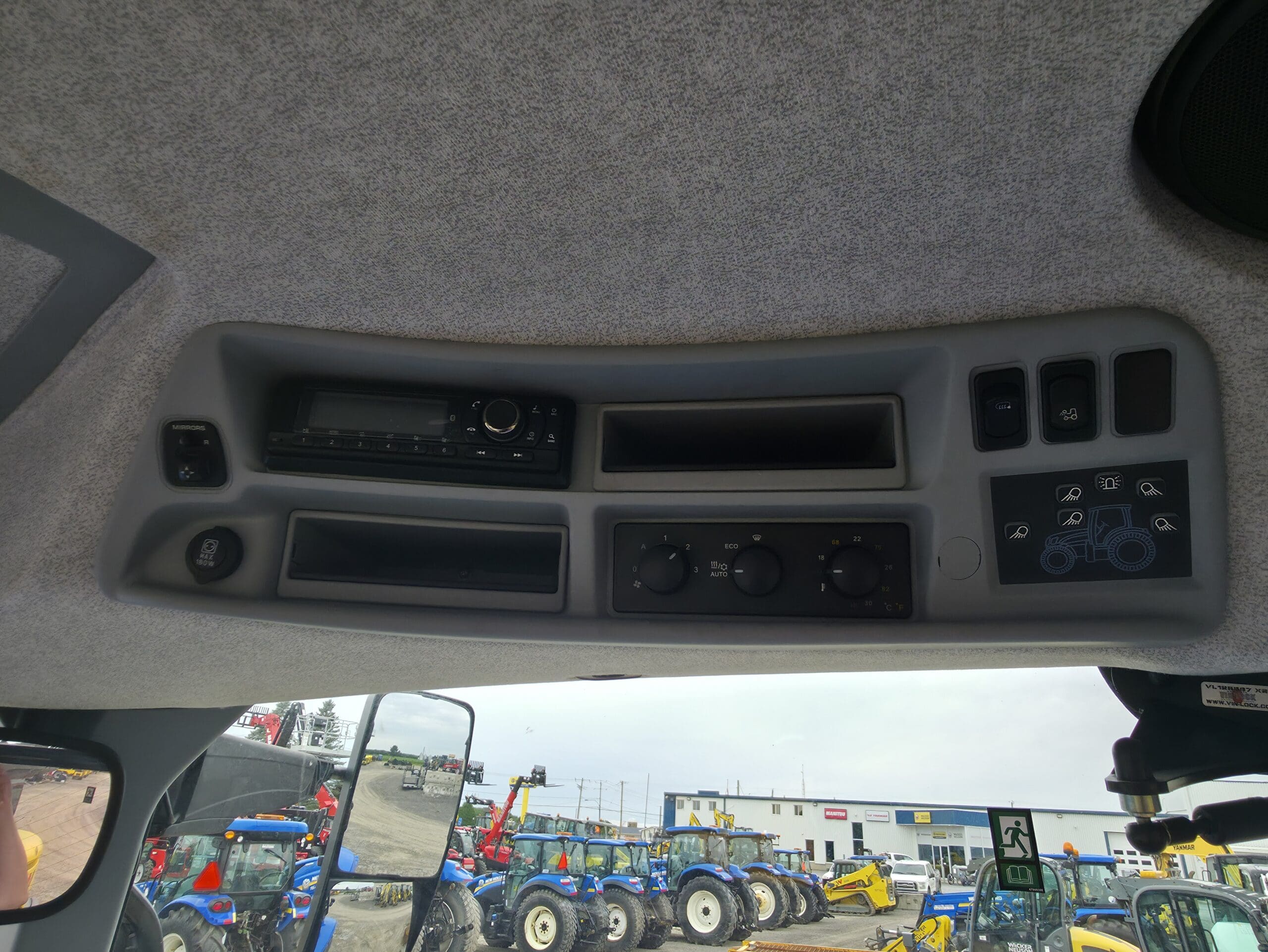 Vue du tableau de commande d'un tracteur avec des boutons, une radio et des compartiments vides, avec divers tracteurs et machines visibles par la fenêtre à l'extérieur.