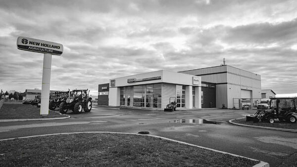 Un concessionnaire agricole New Holland avec des tracteurs garés à l'extérieur sur un terrain pavé sous un ciel nuageux. Le bâtiment principal est doté de grandes fenêtres et d'une enseigne au-dessus de l'entrée.