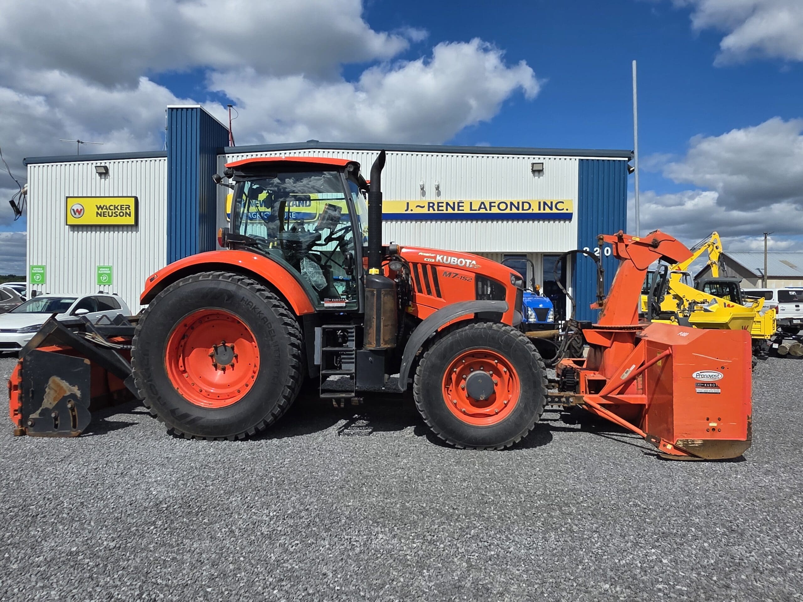 Un grand tracteur Kubota orange avec des accessoires est garé sur du gravier devant un bâtiment industriel bleu et blanc sous un ciel partiellement nuageux. Plusieurs autres véhicules sont visibles à l'arrière-plan.
