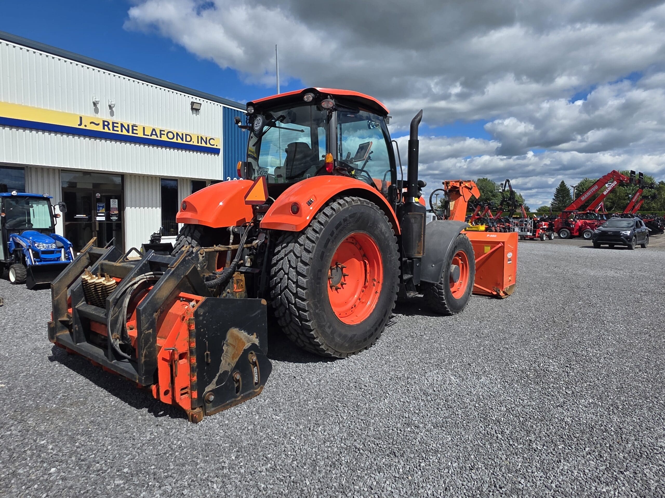 Un gros tracteur orange équipé d'outils avant et arrière robustes est garé sur du gravier à l'extérieur d'un bâtiment portant l'inscription 