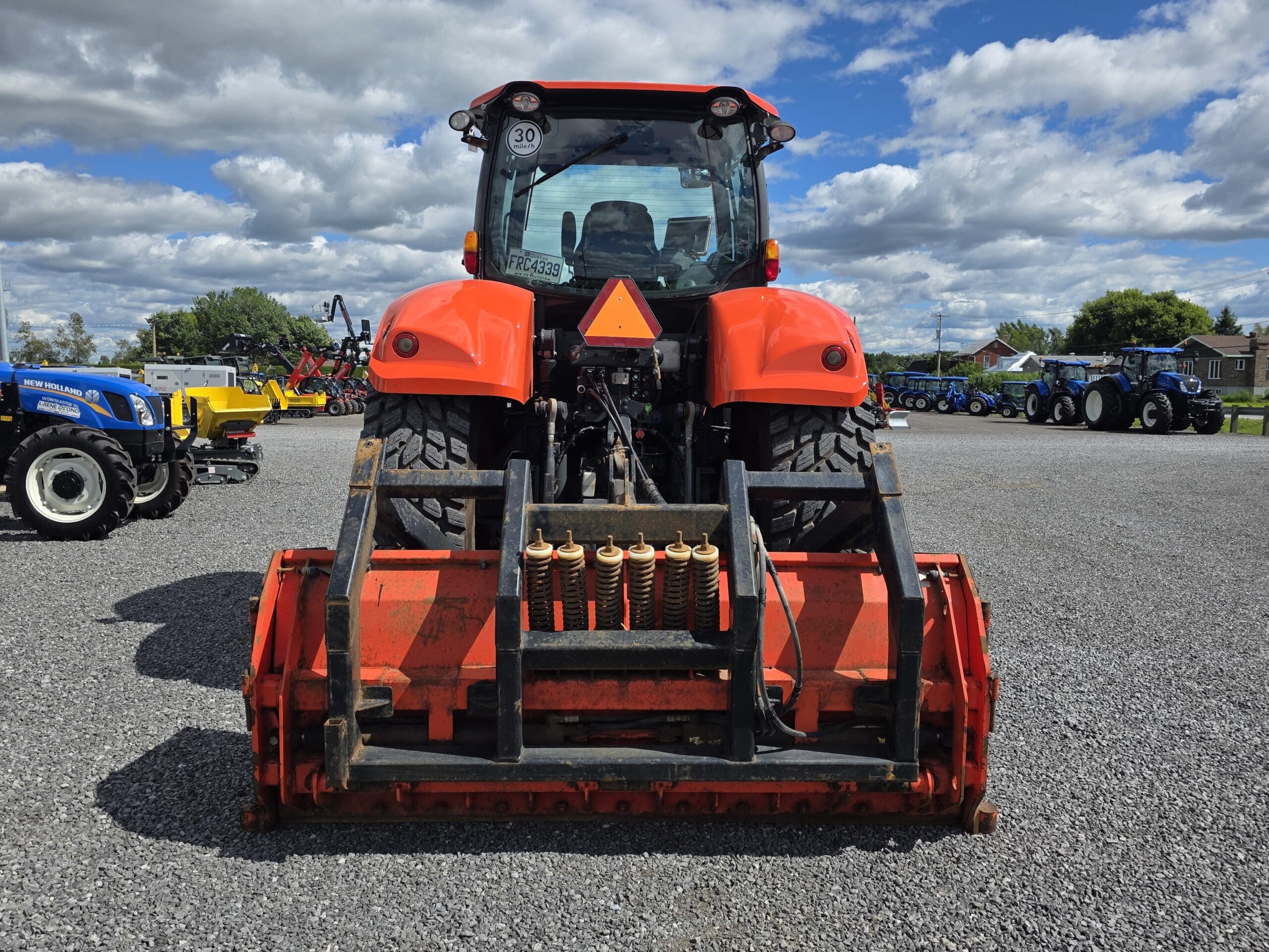 Un tracteur rouge avec un outil frontal est garé sur du gravier, vu de l'arrière. D'autres tracteurs et équipements sont visibles à l'arrière-plan sous un ciel partiellement nuageux.