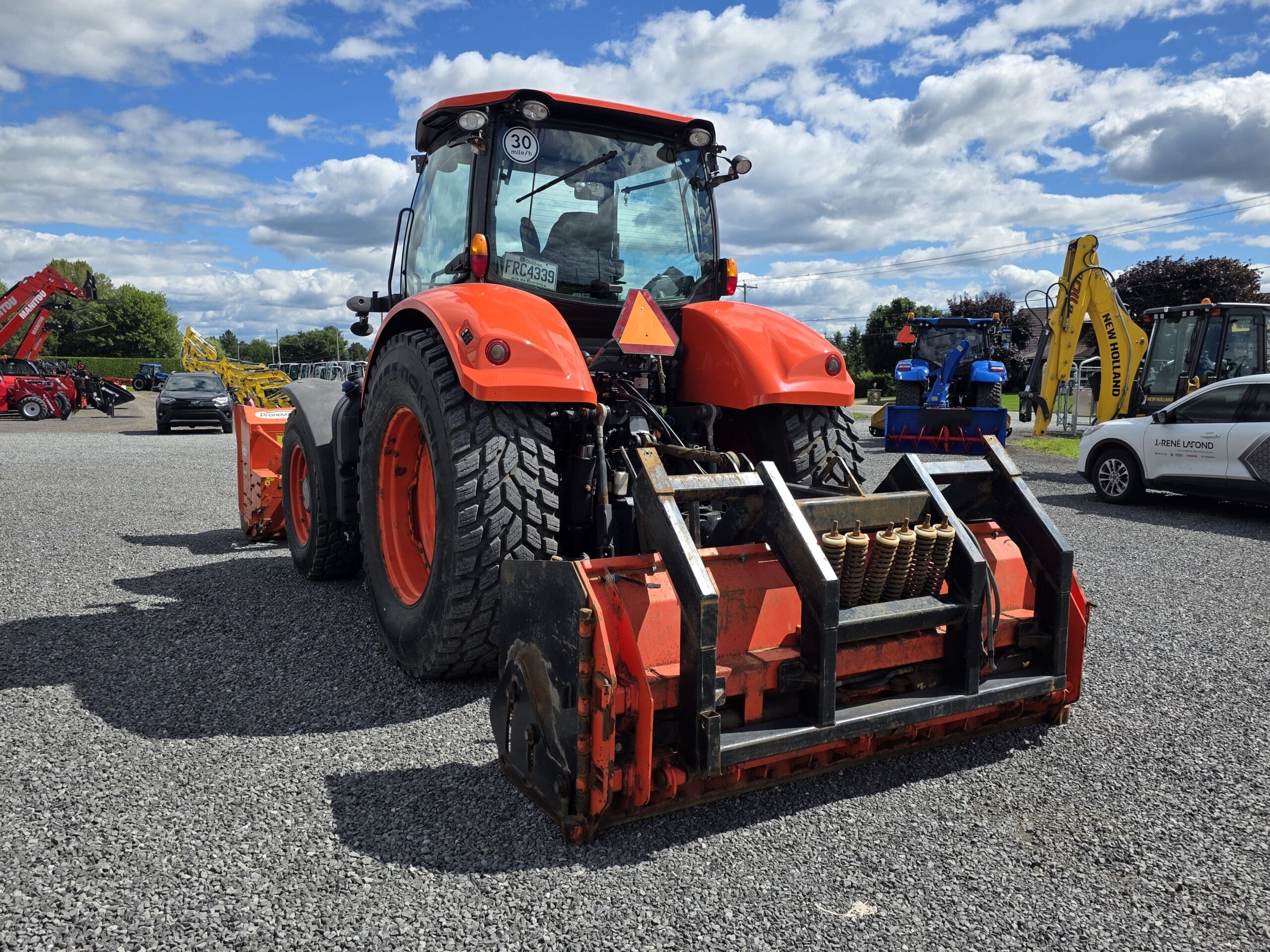 Un tracteur rouge avec de gros pneus arrière et du matériel agricole sur un terrain en gravier, entouré d'autres machines et d'une voiture blanche sous un ciel partiellement nuageux.
