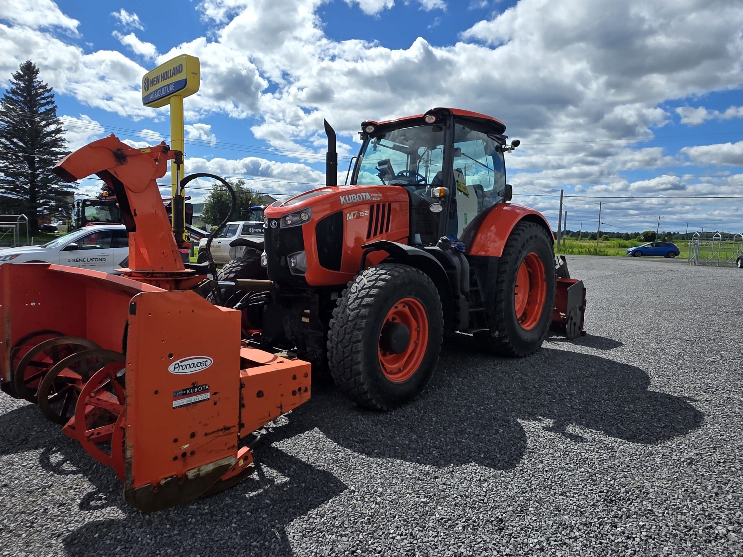 Un grand tracteur Kubota rouge équipé d'une fraise à neige orange montée à l'avant est garé sur du gravier sous un ciel nuageux et ensoleillé. Des voitures et un panneau jaune sont visibles à l'arrière-plan.