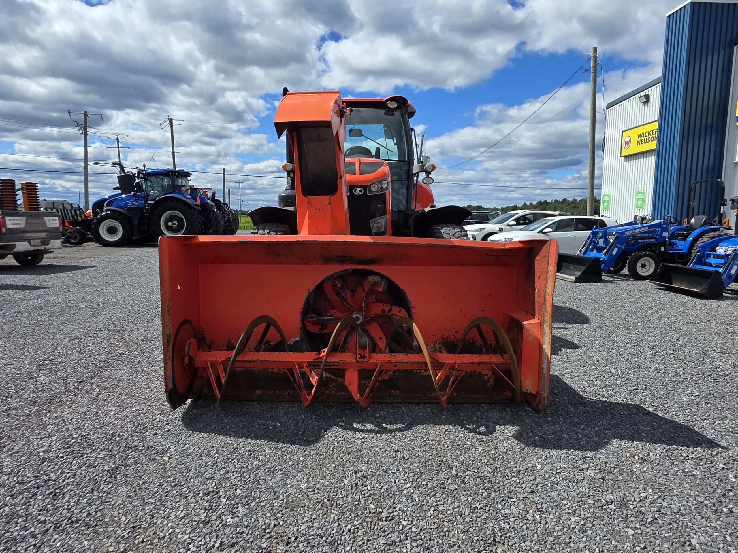Vue de face d'une fraise à neige orange montée sur un tracteur, garé sur du gravier à l'extérieur d'une concession, avec des tracteurs bleus et d'autres véhicules visibles en arrière-plan, sous un ciel partiellement nuageux.